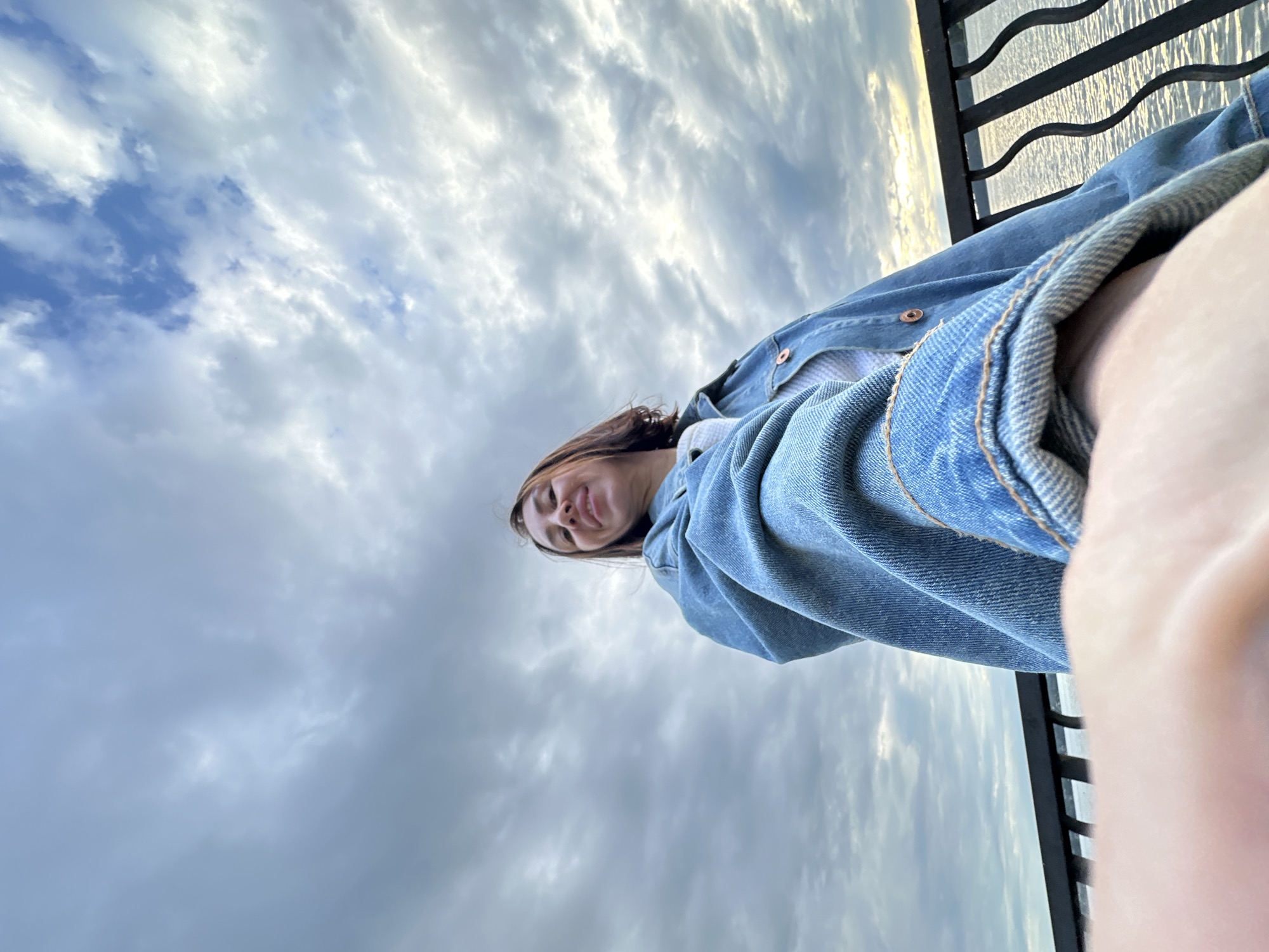 A low-angle, wide shot captures a young woman smiling as she takes a selfie outdoors, with a cloudy sky and a body of water in the background. The woman, with shoulder-length brown hair, wears a denim jacket over a white top. She is positioned in the foreground, leaning slightly forward. A black metal railing runs horizontally across the middle ground, separating her from the rippling water and the overcast sky. The scene appears to be a waterfront area, possibly a promenade or pier, in Votkinsk, Russia. The natural light suggests it is either early morning or late afternoon.