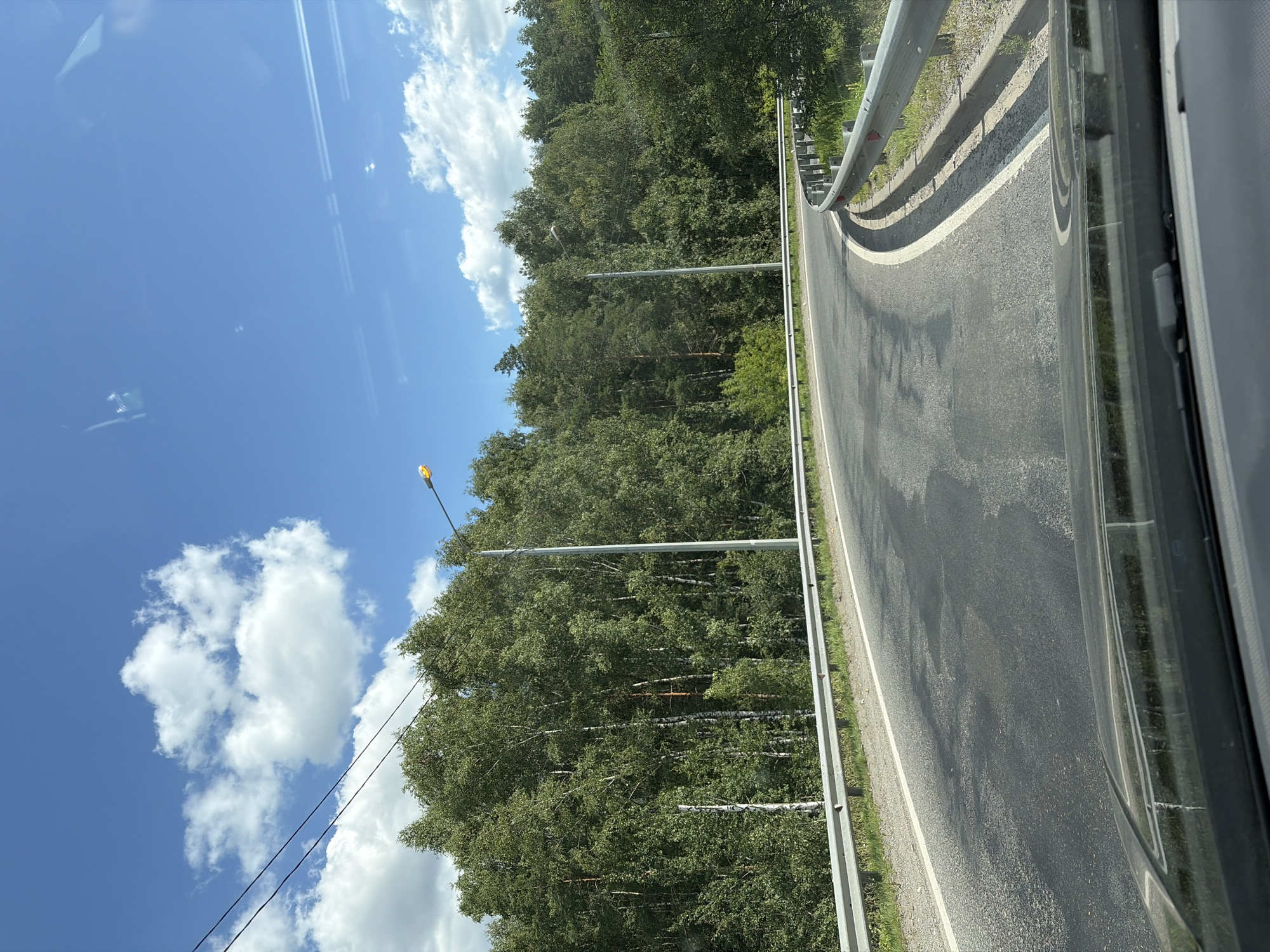 The image depicts a winding asphalt road bordered by a metal guardrail and a dense forest of deciduous trees under a bright blue sky with scattered white clouds. The viewpoint appears to be from inside a vehicle, looking forward through the windshield. There are overhead power lines visible in the upper left portion of the frame. A streetlamp is positioned on the right side of the road. The road surface shows signs of wear and patching. This scene is consistent with a road in a suburban or rural area, such as the outskirts of Noginsk, Russia, characterized by its surrounding green spaces and infrastructure. There are no people or discernible actions taking place.