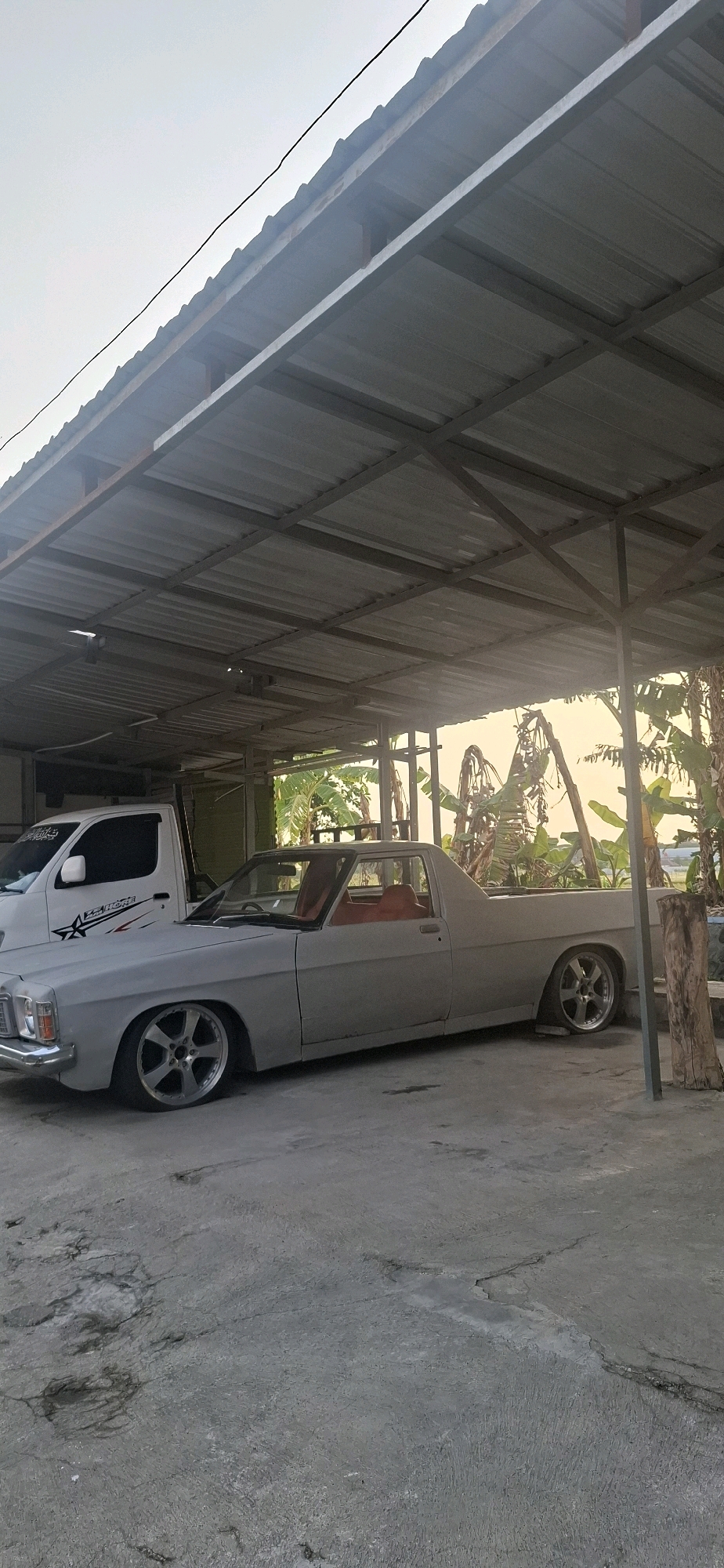 A light gray, vintage pickup truck with a red interior is parked under a corrugated metal roof supported by metal beams. The truck is positioned on a cracked concrete surface. To the left of the truck, a white van with a black and red decal on its side is visible. Behind the truck, banana trees and other foliage are present. The scene appears to be in a covered parking area in Desa Sindon, Indonesia, under a hazy sky. The overall condition of the truck suggests it may be undergoing restoration or modification.