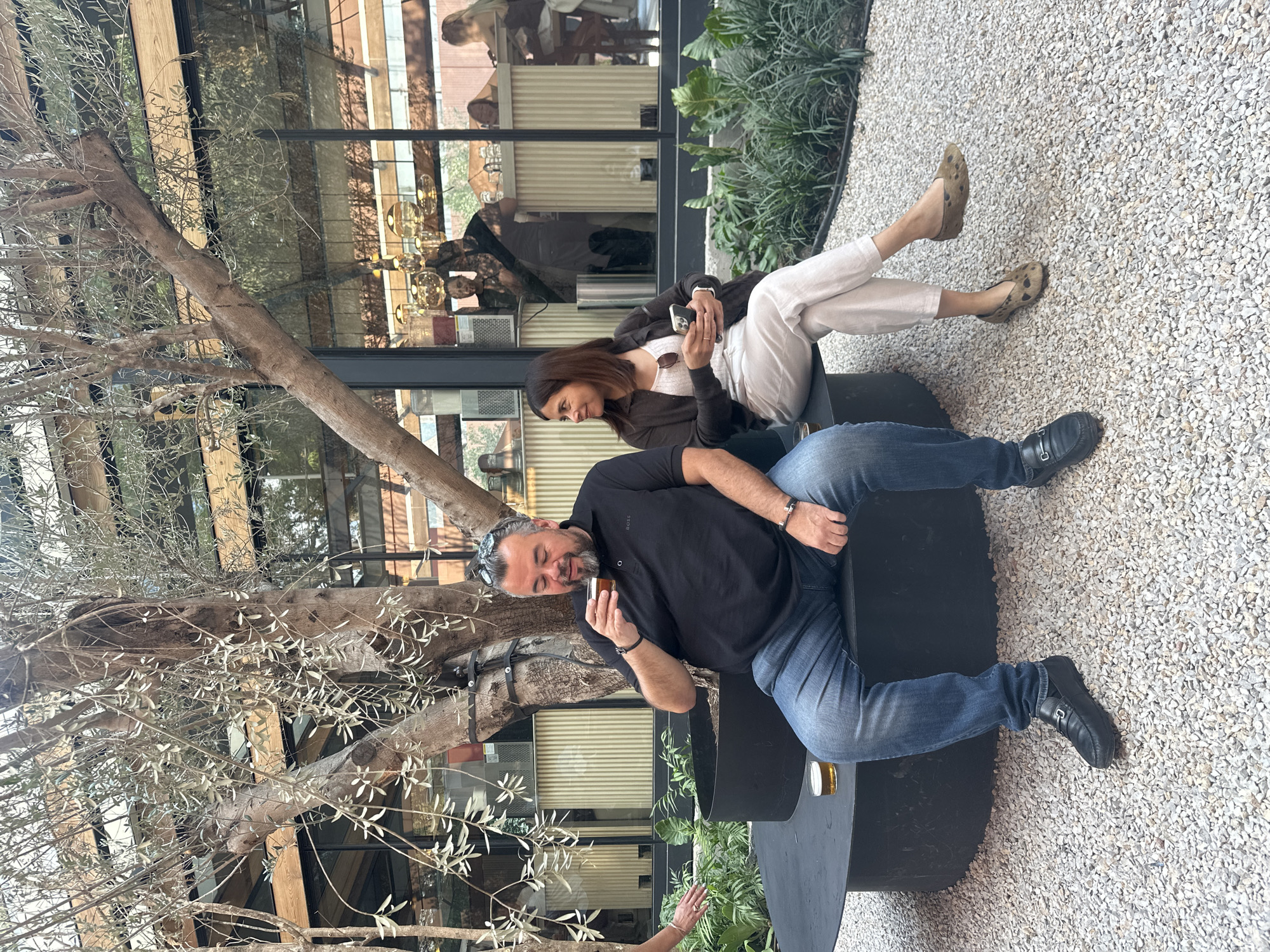 Here is a description of the image:
Two people are seated on a black circular bench in what appears to be an outdoor patio or garden area. The man, with dark hair and a beard, wears a black shirt and blue jeans and holds a glass. The woman, with dark hair, wears a white top, a dark jacket, and light-colored pants and holds a smartphone. The ground is covered with small, light-colored gravel. A tree with bare branches is located beside the bench. A building with large glass windows is in the background, reflecting activity inside.
Based on the style of architecture and landscaping, this could be a patio area in a restaurant or cafe in Mexico City, Mexico.