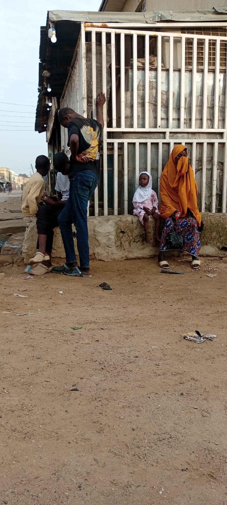 The image captures a street scene in Kano, Nigeria. The foreground is dominated by sandy, dusty ground with some scattered debris. A concrete barrier runs horizontally across the mid-ground, upon which several individuals are positioned. On the left, two young boys are seated, one wearing a yellow long-sleeved shirt and dark pants with beige clogs, the other in a grey t-shirt and black pants. Standing over them is a young man in a black t-shirt with a graphic print and blue jeans, his posture suggesting he is interacting with them. To the right of the concrete barrier, a young girl in a white hijab and pink dress is seated, and next to her, a woman is seated, her face obscured by an orange hijab. She is wearing a colorful patterned skirt. Behind them is a building with a white metal grate and a corrugated metal roof, where several light bulbs are visible, suggesting it might be a shop or stall. The overall environment appears to be an urban setting, possibly a market or roadside commercial area, with a hint of residential buildings in the background. The lighting suggests it is daytime, likely late afternoon given the soft light.