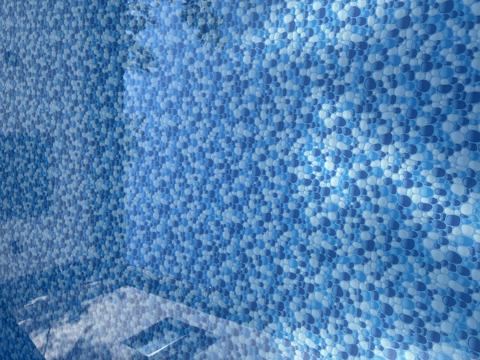 The image displays the blue, pebbled pattern of an in-ground swimming pool liner. The surface is partially covered by a reflection of the sky and surrounding environment, which appears to be a bright, sunny day with white, fluffy clouds. The water's surface is calm, showing clear reflections. There are no people or visible objects within the pool itself. The location is not specified in the image or context.