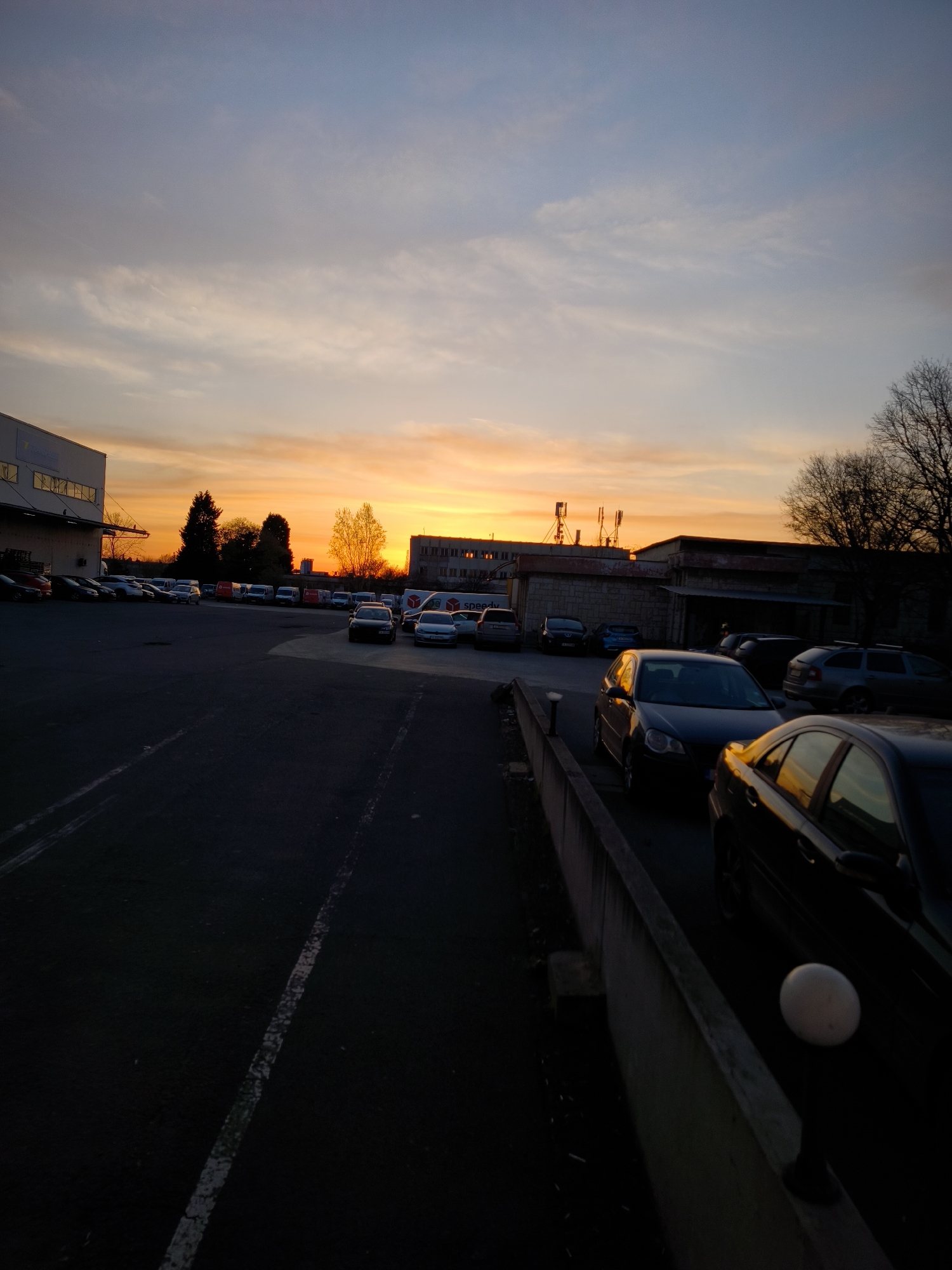 The image depicts a parking lot scene in Burgas, Bulgaria, during sunset. In the foreground, several parked cars are visible along a concrete barrier. A row of cars extends into the middle ground. In the background, buildings are silhouetted against the orange and yellow sky, with telecommunications towers visible on one of the buildings. The sky transitions to blue at the top of the frame. The parking lot pavement is dark. There are a few trees, most without leaves.
