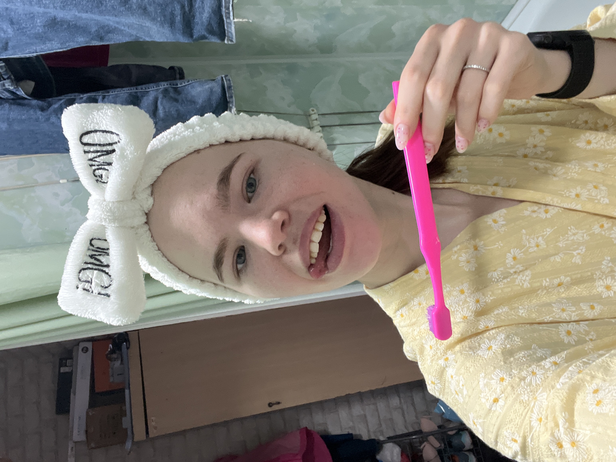 A young Caucasian woman in a yellow daisy-patterned top and a white "OMG" headband is holding a pink toothbrush. She is wearing a ring on her left hand and a black smart watch on her right wrist. She is smiling. The background shows a room with clothes hanging on a rack and shelving. The location appears to be a domestic interior and based on the request, is in the city of Chaikovsky, Russia.