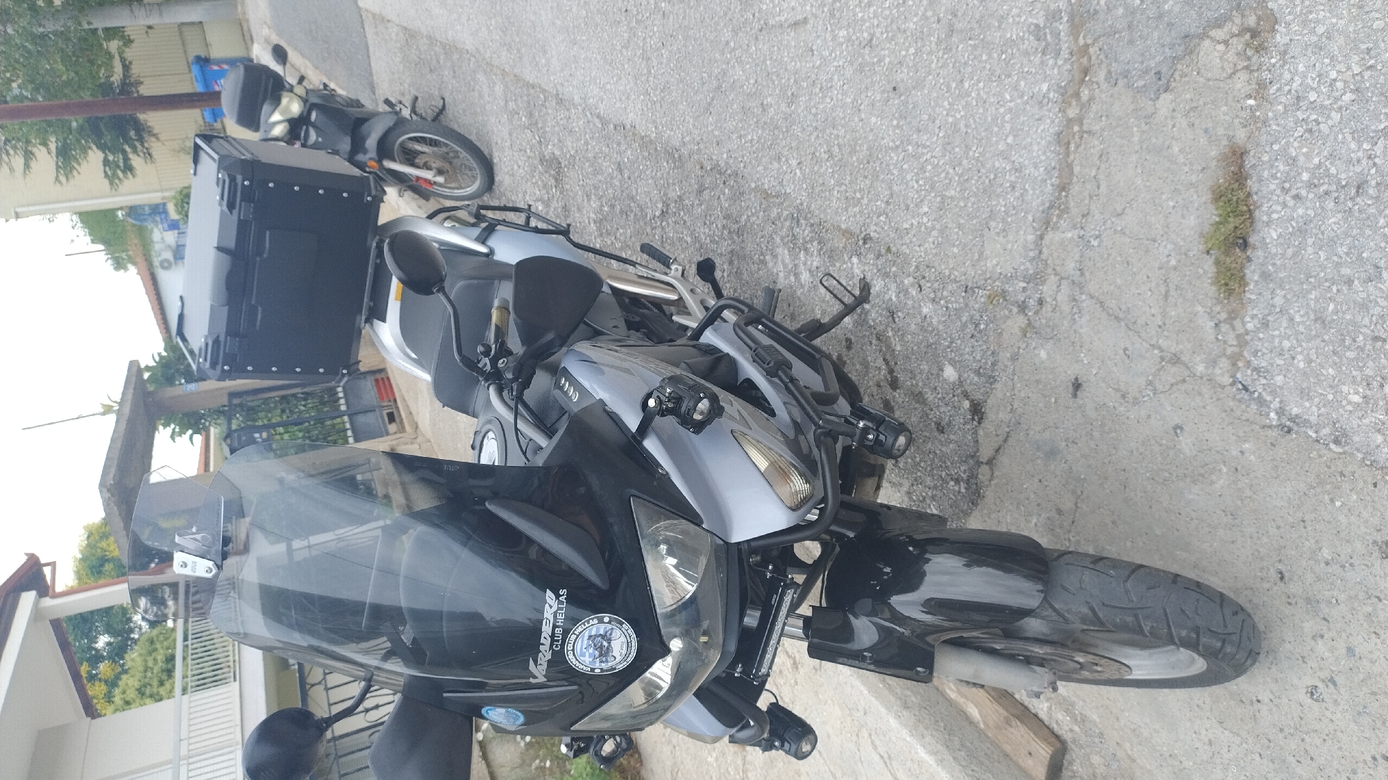 In Νέα Κερασιά, Greece, two motorcycles are parked on a paved street. The primary motorcycle in the foreground is silver and black, with a tall black windscreen, auxiliary lights mounted on the front, and a large black storage box attached to the rear. It has a "Varadero Club Hellas" sticker on the front. A second, smaller black motorcycle is visible behind it, also equipped with a large black storage box. The environment includes a residential building with white railings on the left, a fence, and trees. The ground is a mix of asphalt and cracked concrete, with some sparse vegetation. The sky is overcast.