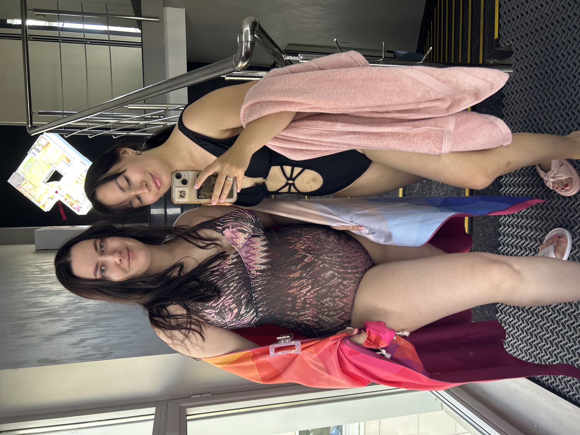 Two women stand in front of a staircase in what appears to be a public building, possibly a sports complex or hotel in Saint Petersburg, Russia. The woman on the left wears a patterned one-piece swimsuit and is draped in a brightly colored sarong. The woman on the right is in a black one-piece swimsuit with cutouts and holds a pink towel. She is taking a selfie with her phone. The environment includes a patterned floor mat, a metal railing, and a window on the left. A digital floor plan is visible on the wall behind the staircase.