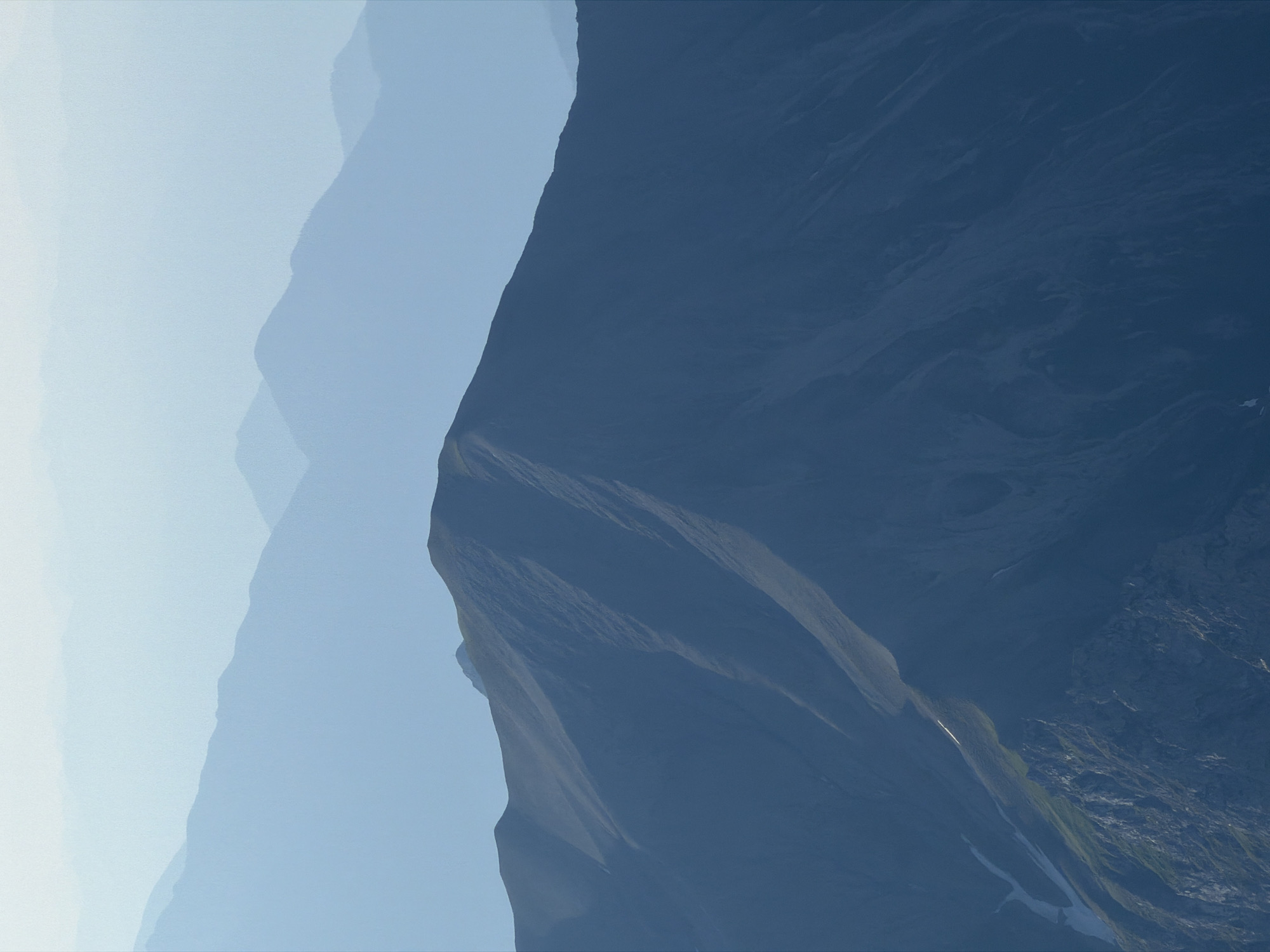 This is a panoramic landscape photograph of layered mountain ranges under a hazy, light blue sky. The foreground features a large, dark blue mountain slope with visible rock formations and patches of green vegetation. To the left, a snow-covered ravine cuts down the mountainside. Further back, several less distinct mountain ranges fade into softer shades of blue due to atmospheric perspective. The overall scene evokes a sense of vastness and natural grandeur, characteristic of alpine environments. There are no people or man-made structures visible in the image, suggesting a remote natural setting. The image is likely a view from a high vantage point, looking out over a chain of mountains.