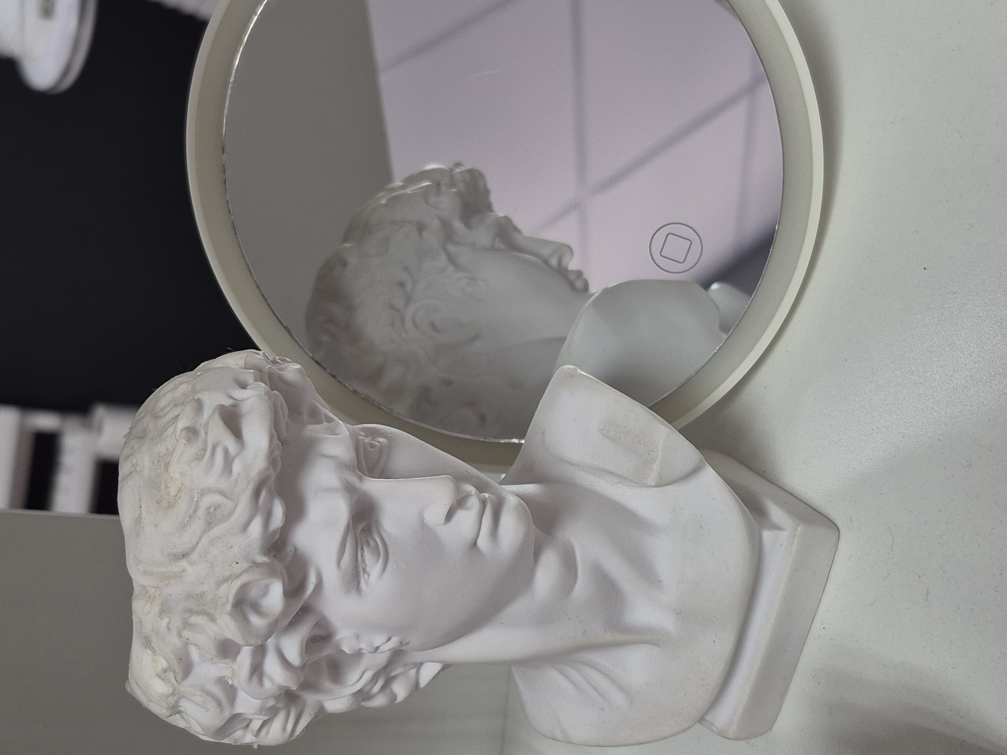 A white bust resembling Michelangelo's David is positioned on the left side of a white surface, angled towards a circular mirror on the right. The bust depicts a male head and shoulders, with curly hair and a thoughtful expression. The mirror reflects a portion of the bust, showing its profile. A small, square icon is visible within the mirror's reflection. The background is blurred, with vertical black and white lines in the upper left and a dark area to the upper right, against a pale, possibly grey or lavender, diffuse background. The scene appears to be indoors, with artificial lighting. There are no people present.