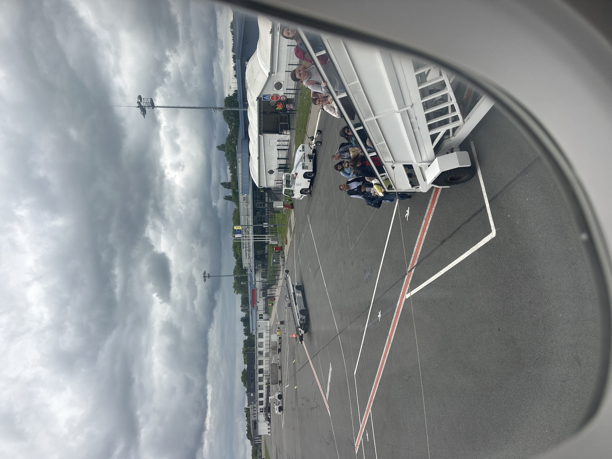 The image captures an airport scene viewed from an aircraft window, partially framed on the right by the window's edge. The sky above is heavily overcast with dark, dense clouds, suggesting gloomy weather conditions.
In the foreground, a white mobile passenger staircase is positioned next to the aircraft, filled with people, likely passengers, who appear to be either boarding or disembarking. Some individuals are standing on the steps, while others are seated on platforms along the staircase.
The tarmac below is asphalt, clearly marked with white and red lines characteristic of airport ground operations. Several ground support vehicles are visible: a white pushback tug is situated near the passenger staircase, and further down the apron, other service vehicles, including a long, low vehicle possibly used for baggage or cargo, can be seen. A person wearing a high-visibility vest is discernible near one of these vehicles, indicating airport staff.
In the background, several airport buildings are visible, including large white tent-like structures, which may be terminal extensions or hangars, and other smaller utility buildings. A tall pole topped with lights or antennae stands prominently against the cloudy sky. Near the buildings, flags on poles are faintly visible, with one resembling the European Union flag. Visible text includes a red "STOP" sign and a blue sign with a white arrow near the buildings, and ground markings like "2" and "3" on the tarmac.
The scene depicts a typical airport operation, likely a passenger transfer, during daylight hours under an overcast sky. The location context is Tillé, France.