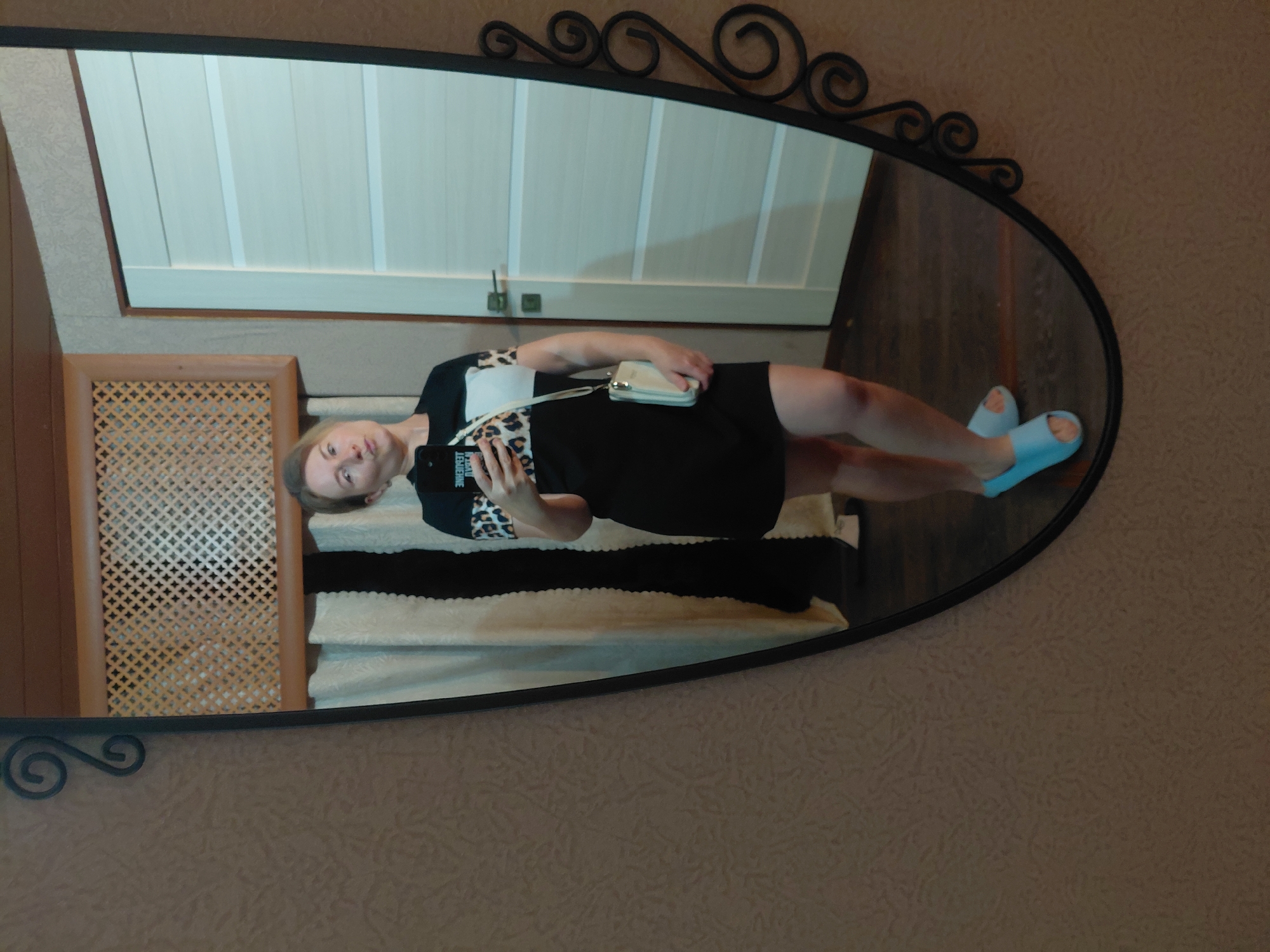 A woman is taking a mirror selfie in a room. She is wearing a black dress with leopard print accents on the sleeves and a white panel on the chest. She is also carrying a light-colored clutch bag with a strap. She has light blue slip-on sandals on her feet. The mirror is framed by an ornate black metal border and reflects a doorway with white doors on the right side of the image, and curtains and a wooden lattice on the left. The walls of the room are covered in textured wallpaper. The floor appears to be dark wood. The woman is in a room, possibly a home, in Novosibirsk, Russia.