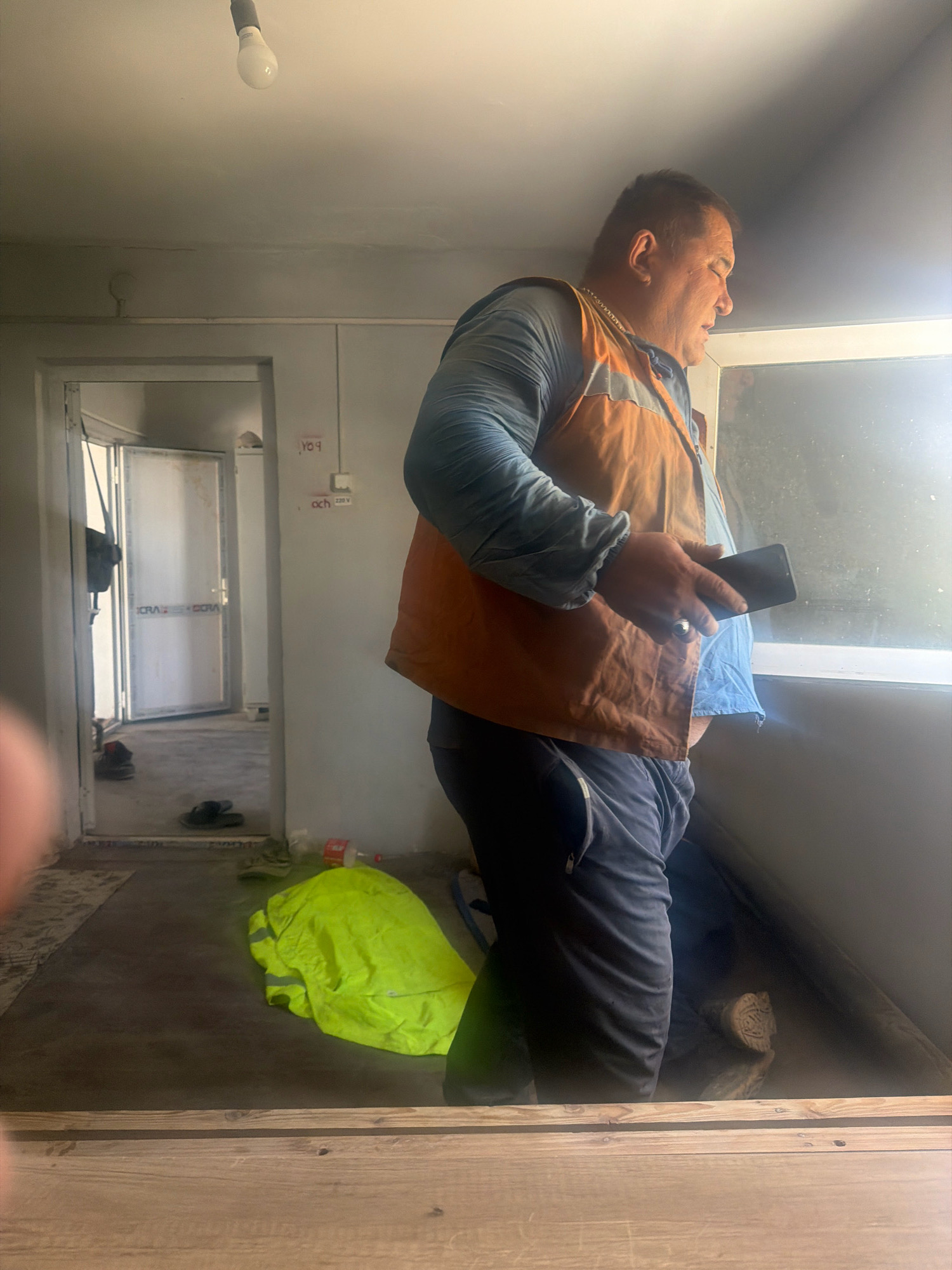 The image depicts an interior scene, possibly a temporary structure or room in Besapan, Uzbekistan. In the foreground, a wooden surface is visible. The mid-ground is dominated by a man standing near a window, looking out. He is wearing a long-sleeved blue shirt and an orange reflective vest over it. He is holding a dark smartphone in his right hand. His left hand is resting on his stomach area. The room has light-colored walls. An open doorway to the left reveals a hallway or another room with a door at the end. On the floor near the doorway, a pair of dark slippers is visible. Further into the room, a bright yellow reflective jacket is folded on the floor, next to a red bottle. The ceiling has a light fixture with a bulb. The overall impression is of a utilitarian space, possibly a worksite or a temporary accommodation. The lighting suggests it is daytime, with sunlight streaming in from the window. There is some writing on the wall above the doorway, including characters that appear to be Cyrillic.