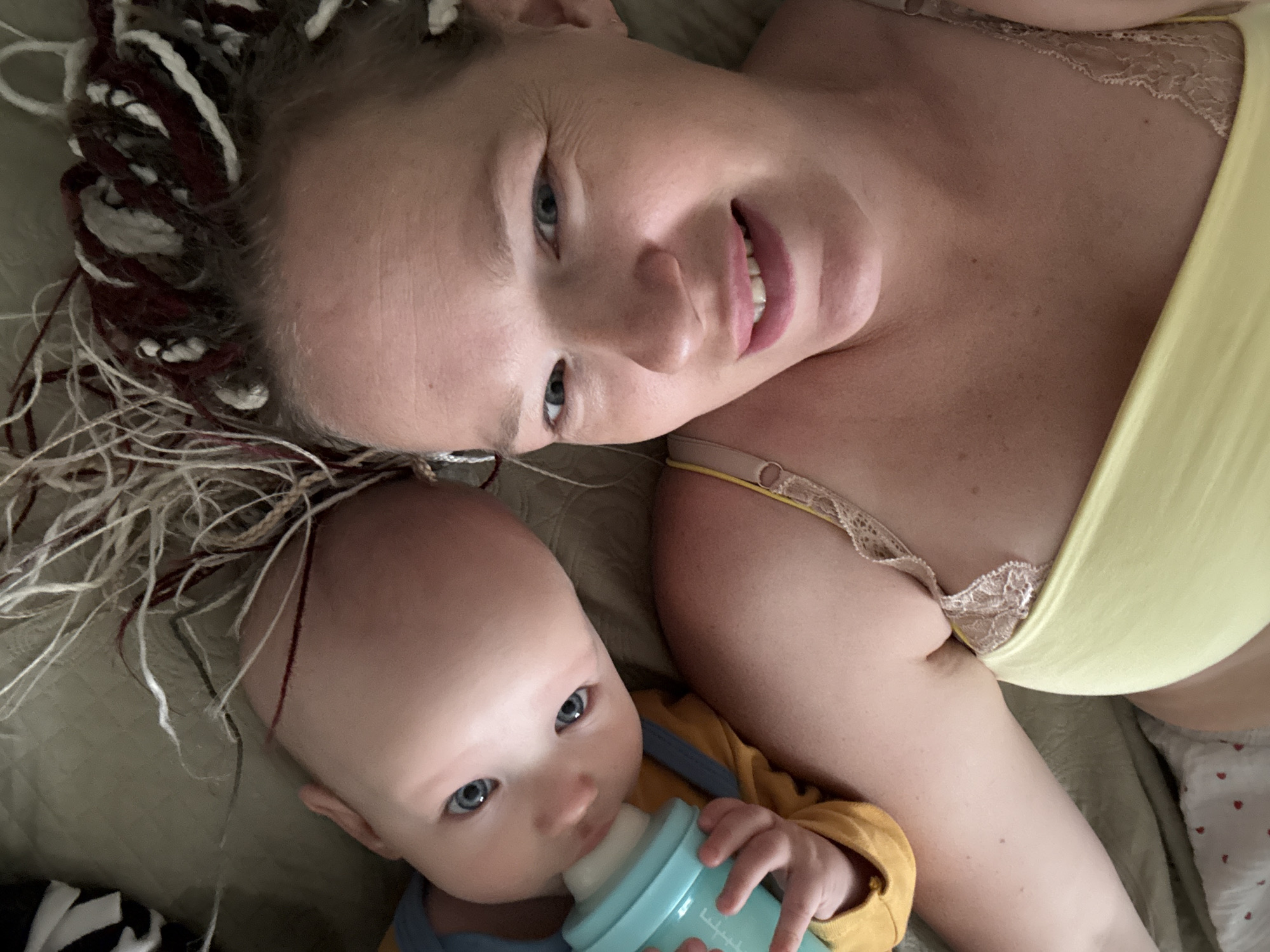 The image shows a close-up of a woman and a baby lying down, with their heads angled towards the camera. The woman, wearing a yellow top with lace trim, is positioned on the left side of the frame, and her face is visible upside down. She has light-colored eyes and pink lipstick. The baby, on the right side of the frame, is also upside down and is holding and drinking from a turquoise baby bottle. The baby has light-colored eyes and is wearing a yellow long-sleeved shirt. Their hair appears to be a mix of blonde and brown, with some lighter streaks. They are lying on a textured surface, possibly a bed or couch, which appears to be a muted green color. The overall lighting suggests an indoor setting. There is no indication of the city of Бордуки, Russia in the image.