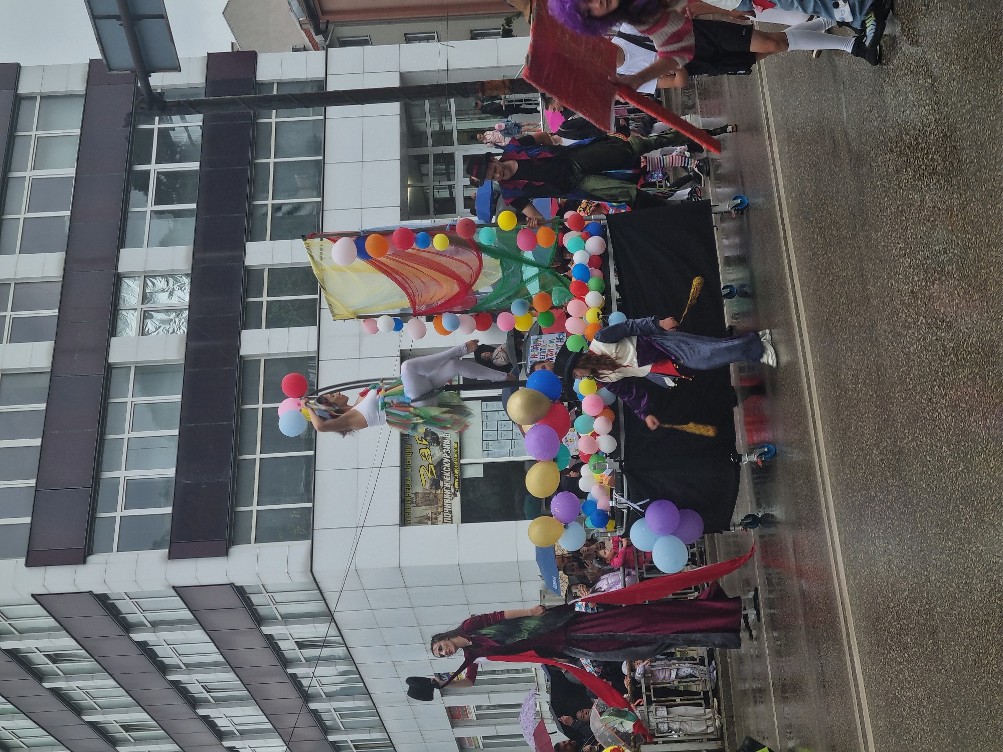 A street-level photograph shows a parade in Gabrovo, Bulgaria. The parade includes floats, people in costume, and balloons. There is a modern building in the background with a grid-like facade. It appears to be a cloudy, overcast day, and the street is wet, suggesting it might have been raining. People are watching the parade. The float in view is decorated with colorful balloons, and performers are on top of it. One performer appears to be suspended in the air, possibly using wires or a harness. People on the float are dressed in costumes and appear to be part of a performance. The street seems to be lined with spectators observing the procession.