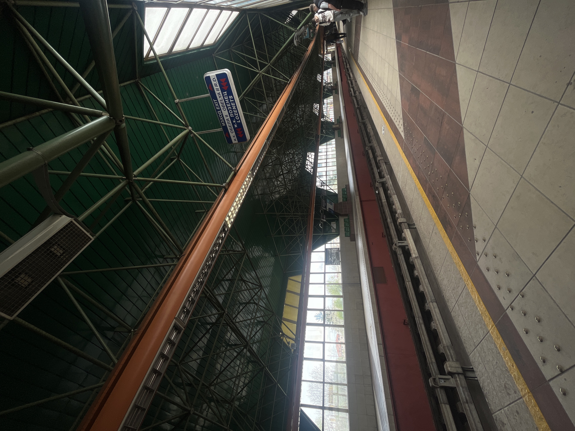 The image shows the interior of a Sofia Metro station in Bulgaria. The view is from a high angle, likely from the upper level of the station, looking down at the platform. The platform itself has a tile floor with a reddish-brown section closer to the tracks and a lighter gray section further away. A yellow safety line runs along the platform edge. Above the platform, there is a complex network of green steel structural beams and supports, some of which run diagonally. Along the center of the image is an orange structure. Windows along the top and side allow natural light to enter. A sign in Bulgarian and English hangs from the structure, indicating "Exit" toward "zh.k. Obelya II R.D." and "zh.k. Lozenets." Some people are visible standing on the platform.