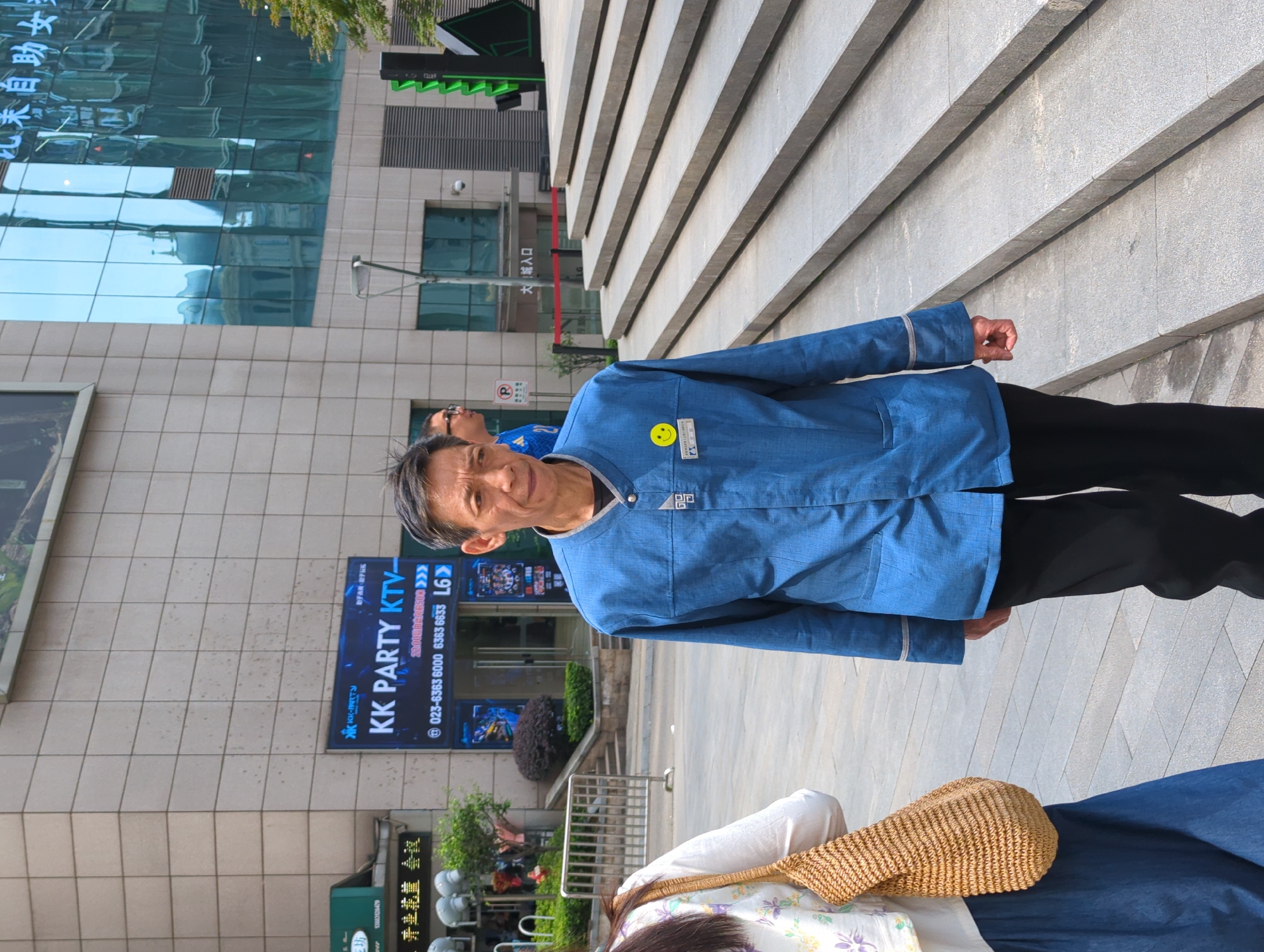 An eye-level outdoor shot depicts an elderly Asian man wearing a blue button-down shirt and black pants, standing on light gray concrete steps. He is looking towards the camera with a slight smile. On his shirt is a small badge with a yellow smiley face and what appears to be a number. Partially visible at the bottom of the frame are a woman's floral-patterned white top and a brown woven bag, along with a portion of what appears to be a blue dress.
Behind the man is a commercial building with a glass facade reflecting other buildings and the sky. To the left of the man, a storefront displays the words "KK PARTY KTV" in large blue letters. A street sign below reads "023-6363 6000 6363 6633 L6." Various storefront signs in Chinese characters are also visible, along with decorative plants and a metal fence.
The location is implied to be Yuzhong District, China, based on the telephone area code 023 in the signage.