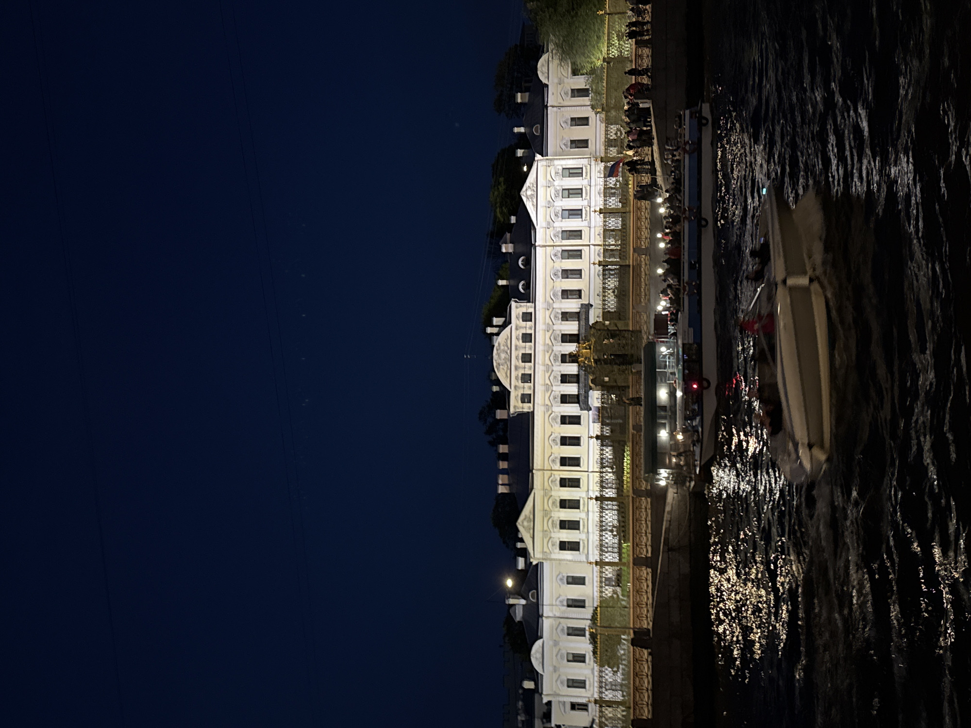 Here is a description of the image:
The image shows a night scene in Saint Petersburg, Russia. A white, multi-story building with many windows is illuminated and stands along a riverbank. The building appears to be a palace or historical structure. A metal fence runs along the riverbank, separating the building from a walkway where people are gathered. Reflections of lights shimmer on the surface of the water. A boat is visible in the water, close to the viewer. The left side of the image is dominated by a dark blue night sky.