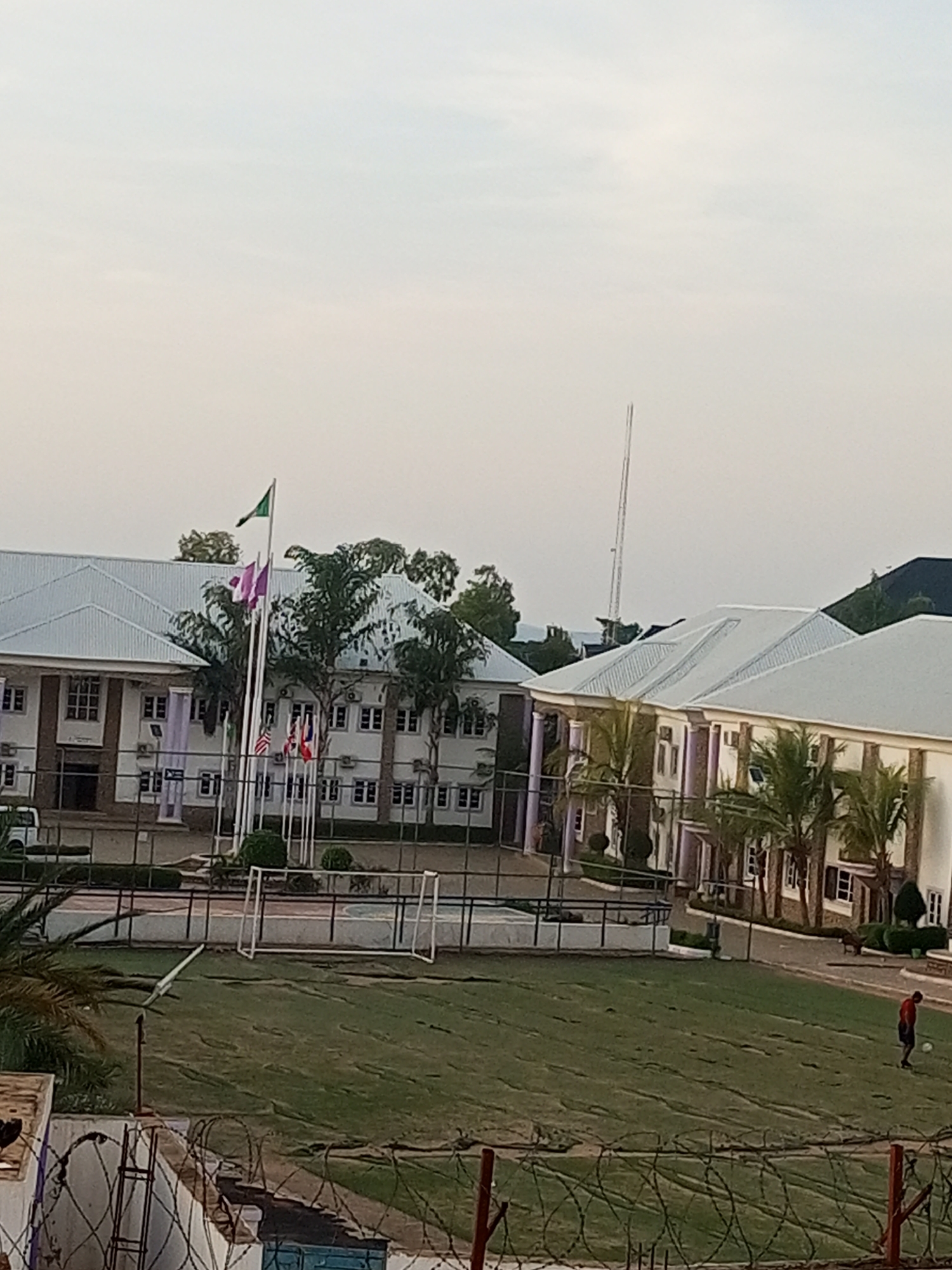 The image depicts a school campus in Gombe, Nigeria. The foreground features a large grassy field with a soccer goal and a person wearing a red shirt and black shorts playing soccer. In the mid-ground, there are several buildings with white, gabled roofs and multiple windows. Flags are flying on flagpoles in front of one of the buildings, including what appears to be the Nigerian flag and other national flags. There are also palm trees and other vegetation around the buildings. The background shows a clear sky and a distant view of hills. A tall, slender tower, possibly a communication tower, is visible in the distance. The overall impression is of a well-maintained educational institution.