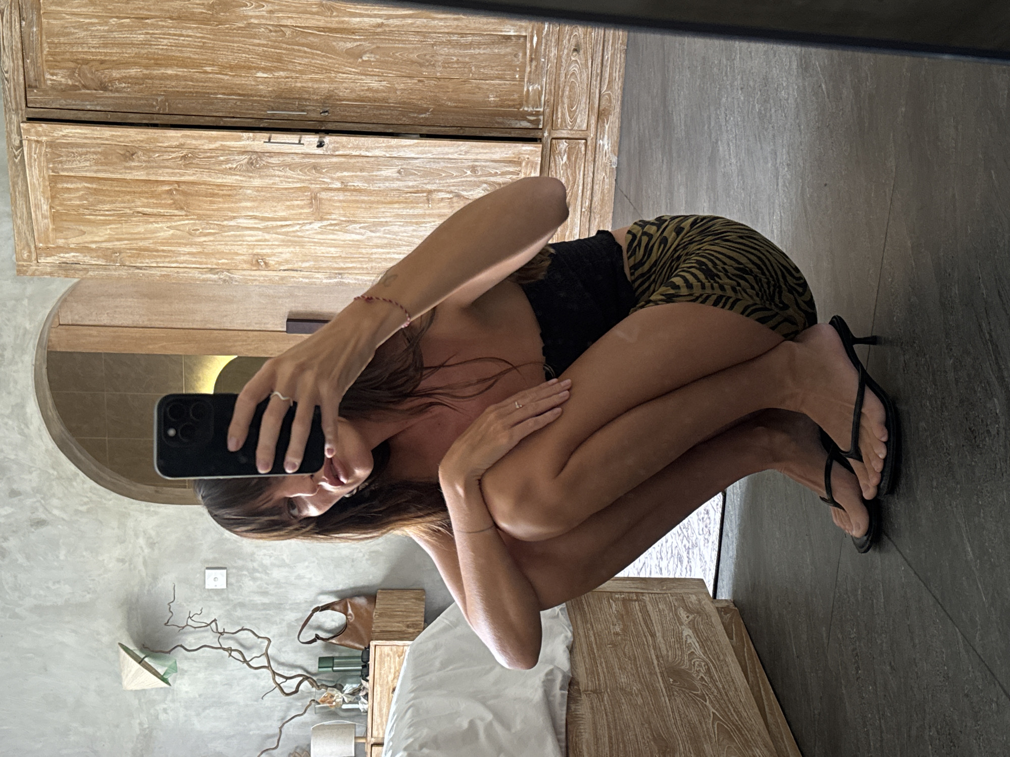 The image captures a woman taking a mirror selfie while squatting in an indoor room, likely in North Kuta, Indonesia. She has long brown hair and is wearing a black top paired with a green and black tiger-print mini skirt. On her feet are black high-heeled flip-flops. She holds a black smartphone towards her reflection in a large mirror, where her face is visible playfully sticking out her tongue. A red string bracelet is on her left wrist, and a ring adorns her right ring finger.
The room features a rustic aesthetic with prominent light-colored, heavily-grained wooden furniture, including a large wardrobe or cabinet with a slightly ajar upper compartment, and a bedside table next to a bed partially covered with white sheets. The walls appear to be light grey concrete or stucco, contrasting with a darker grey tiled or polished concrete floor. Decorative elements include a dried, twisted branch arrangement on the bedside table and a small, white conical hat-shaped ornament hanging on the wall. A brown bag and some bottles are also visible on the bedside table. The overall setting suggests a modern yet natural resort or hotel environment.