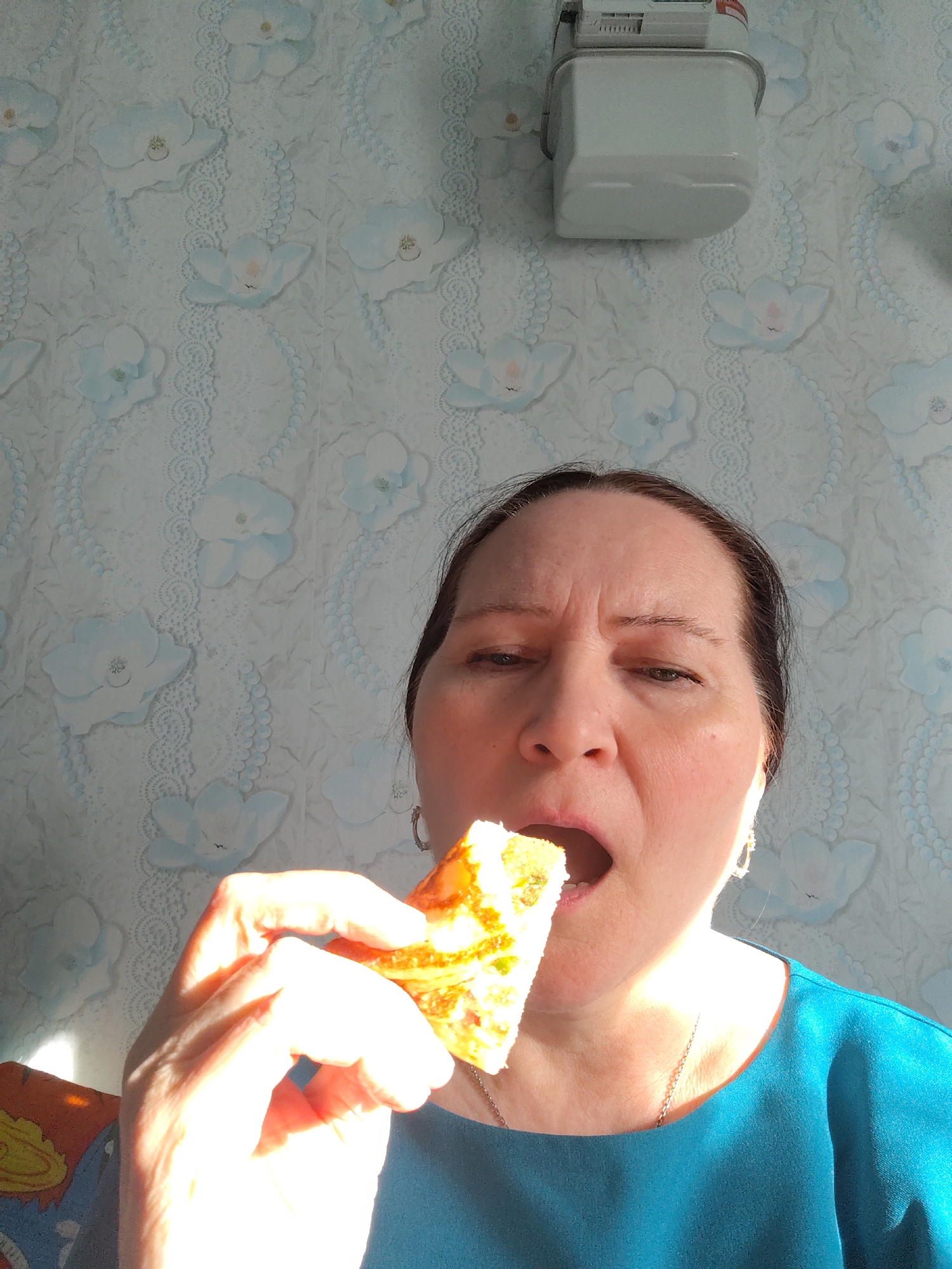 A medium close-up shot captures a woman in the act of eating indoors. The woman, appearing middle-aged with dark hair pulled back, is positioned centrally, looking upwards with her mouth open as she holds a piece of golden-brown food (resembling pie or a savory pastry) in her right hand, poised to eat. Her forehead is slightly furrowed, indicating concentration. She wears a teal-colored top and a thin necklace.
Bright, direct sunlight illuminates the left side of her face, neck, and hands, creating strong highlights and suggesting it is daytime, possibly morning or afternoon. The background features a light-toned wallpaper with a pattern of light blue flowers and vertical lace or beaded designs. In the upper right, a white utility box, likely a gas meter, is mounted on the wall. Some faint, unreadable text is visible on the meter. The scene depicts a candid, everyday moment within a home.