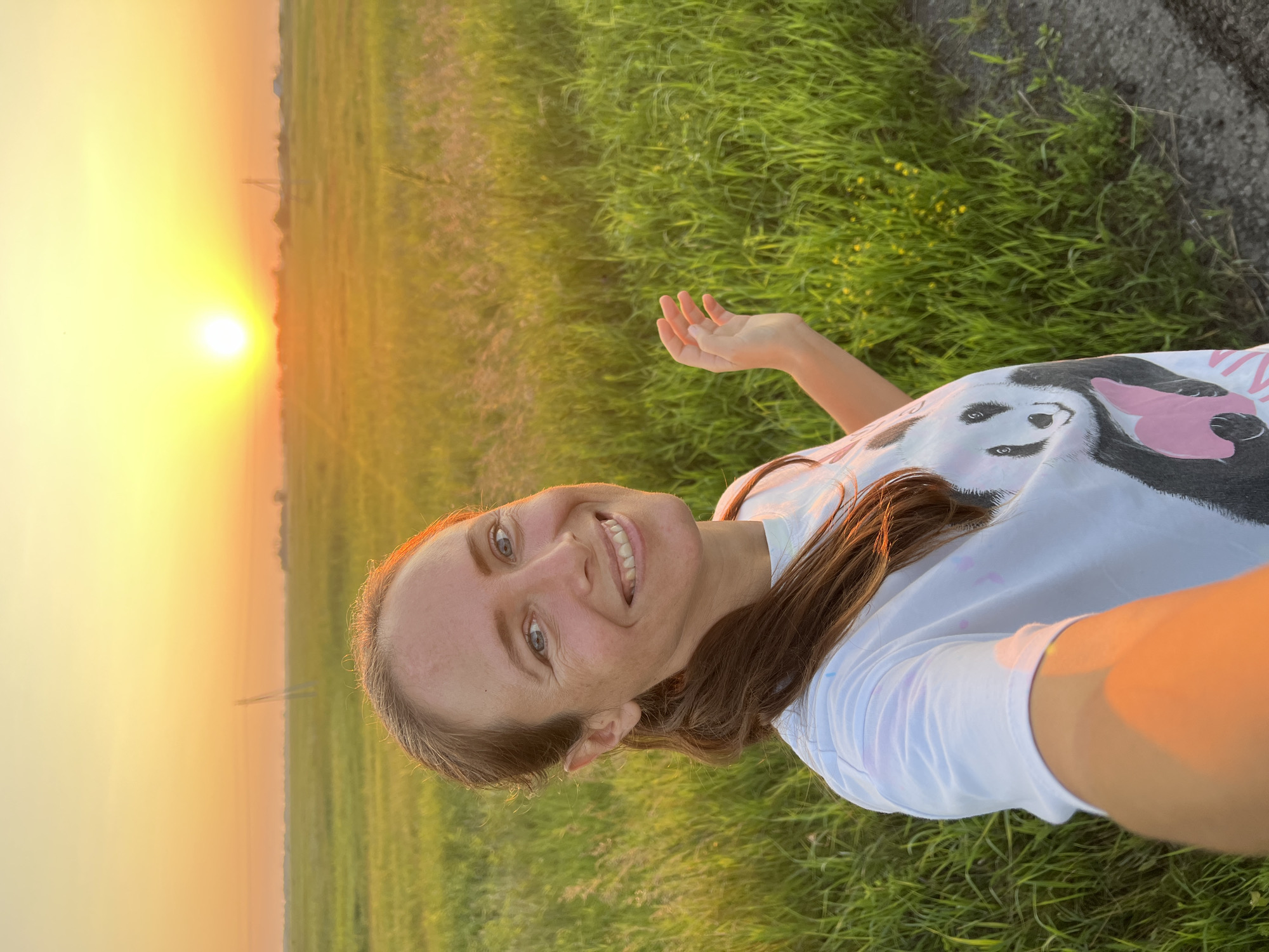 A woman with brown hair pulled back in a ponytail smiles at the camera, taking a selfie outdoors at sunset. She wears a white t-shirt with a panda graphic. Her right arm is extended to the side, with her hand open. The background is a field of green grass, with a bright orange sunset in the distance. Utility poles are visible on the horizon. The scene appears to be in a rural area.