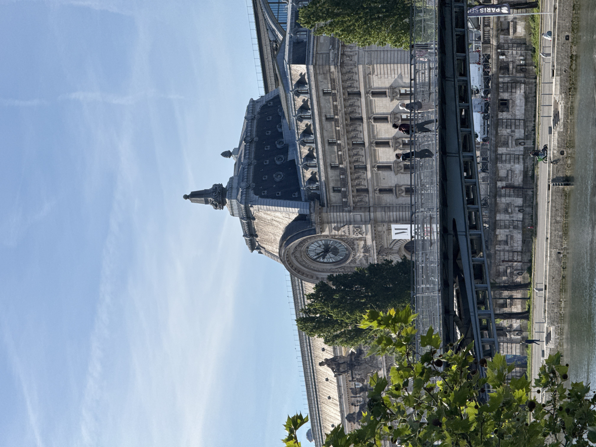 The image captures a partial view of the Musée d'Orsay in Paris, France, from a slightly low angle. The museum's distinctive architecture is visible, including its clock tower and ornate façade. A bridge spans the River Seine alongside the museum, with people walking and cycling along the riverbank below. Some trees and foliage are visible in the foreground, partially obscuring the view. The sky is clear and blue.