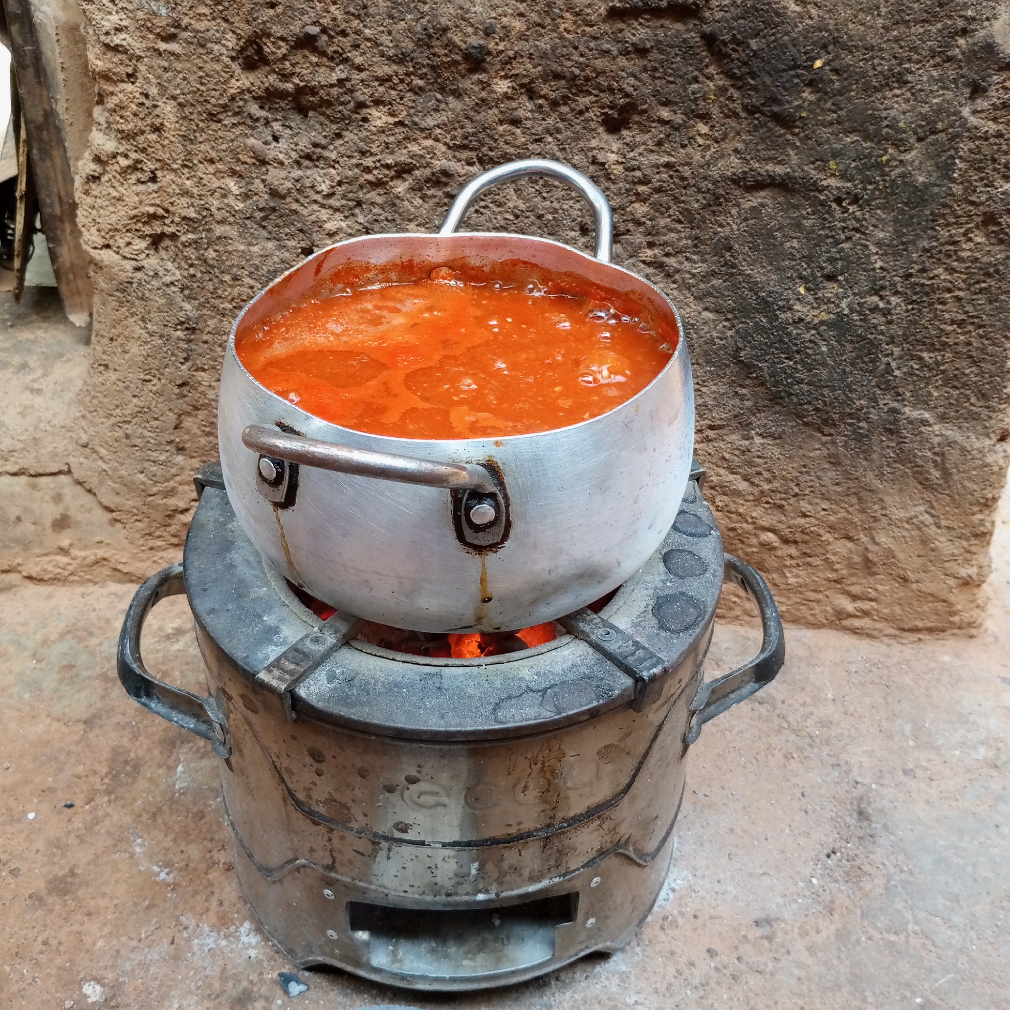 A silver cooking pot filled with a vibrant orange liquid, possibly stew or soup, sits atop a charcoal stove with glowing embers visible inside. The stove has two handles and shows signs of wear and use. The setting appears to be outdoors, with a rough, textured wall in the background, potentially a building constructed with locally sourced materials. This scene is likely located in the city of Jos, Nigeria, based on the materials and cooking setup.