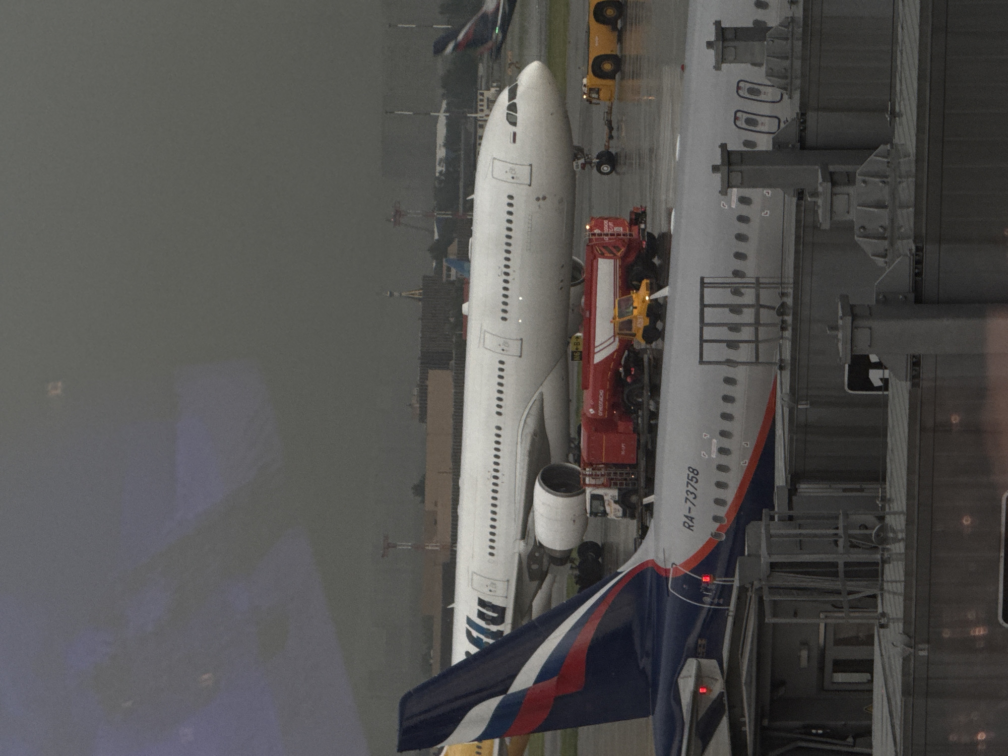 The image shows an airport tarmac scene at Sheremetyevo International Airport in Khimki, Russia. In the foreground, the tail of a Russian flag-themed airplane is visible, with the registration "RA-73758" clearly marked on the fuselage of the plane behind it. A red fuel truck is parked next to the plane, and a yellow tow tractor is attached to the nose wheel of a large white passenger airplane in the background. The ground is wet, indicating recent rain. The sky is overcast and appears dark and stormy. In the distance, airport infrastructure, including control towers and other aircraft, can be seen. The scene suggests a typical operational day at a busy international airport, possibly during inclement weather.