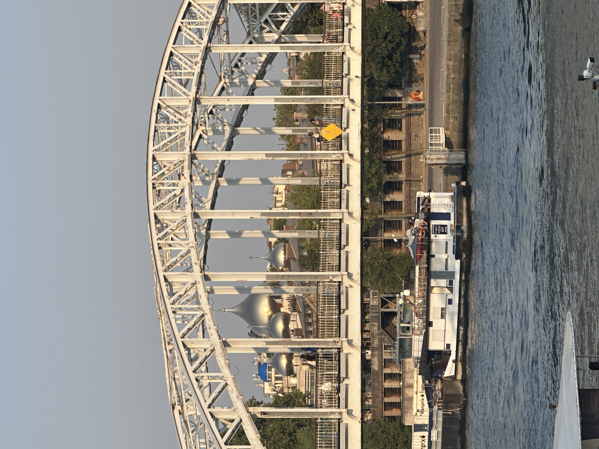 The image shows a view of the Pont de Bir-Hakeim, a bridge crossing the Seine River in Paris, France. The bridge has a distinctive arched metal structure. In the background, visible through the bridge's structure, are several domed buildings, likely part of a church or cathedral, one of which has a bright blue dome. The bridge itself has a pedestrian walkway with a railing, and several people can be seen walking on it. A yellow diamond-shaped sign is attached to the railing of the bridge. Below the bridge, on the river, a white barge or boat is docked. The water of the Seine is visible in the foreground, and the riverbank with buildings and trees is visible behind the barge. The sky is clear and a pale blue, suggesting it is daytime, possibly late afternoon due to the warm light.