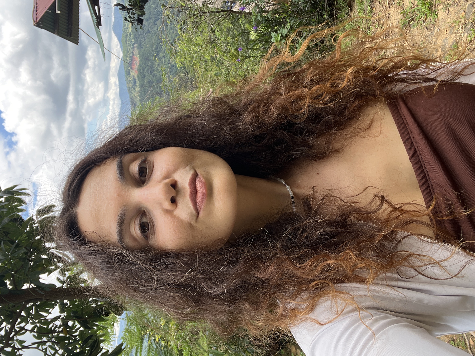 A medium shot portrait of a woman with long, curly brown hair, wearing a white open shirt over a brown top, is captured outdoors. She has fair skin and is looking towards the camera with a neutral expression. A pearl necklace is visible around her neck. The background reveals a lush green landscape with rolling hills and trees, under a cloudy sky. A portion of a building with a red roof is visible in the upper right corner, suggesting a rural or mountainous setting, possibly in the city of Sơn Điền, Vietnam. The ground in the foreground is covered with dirt and small patches of grass.