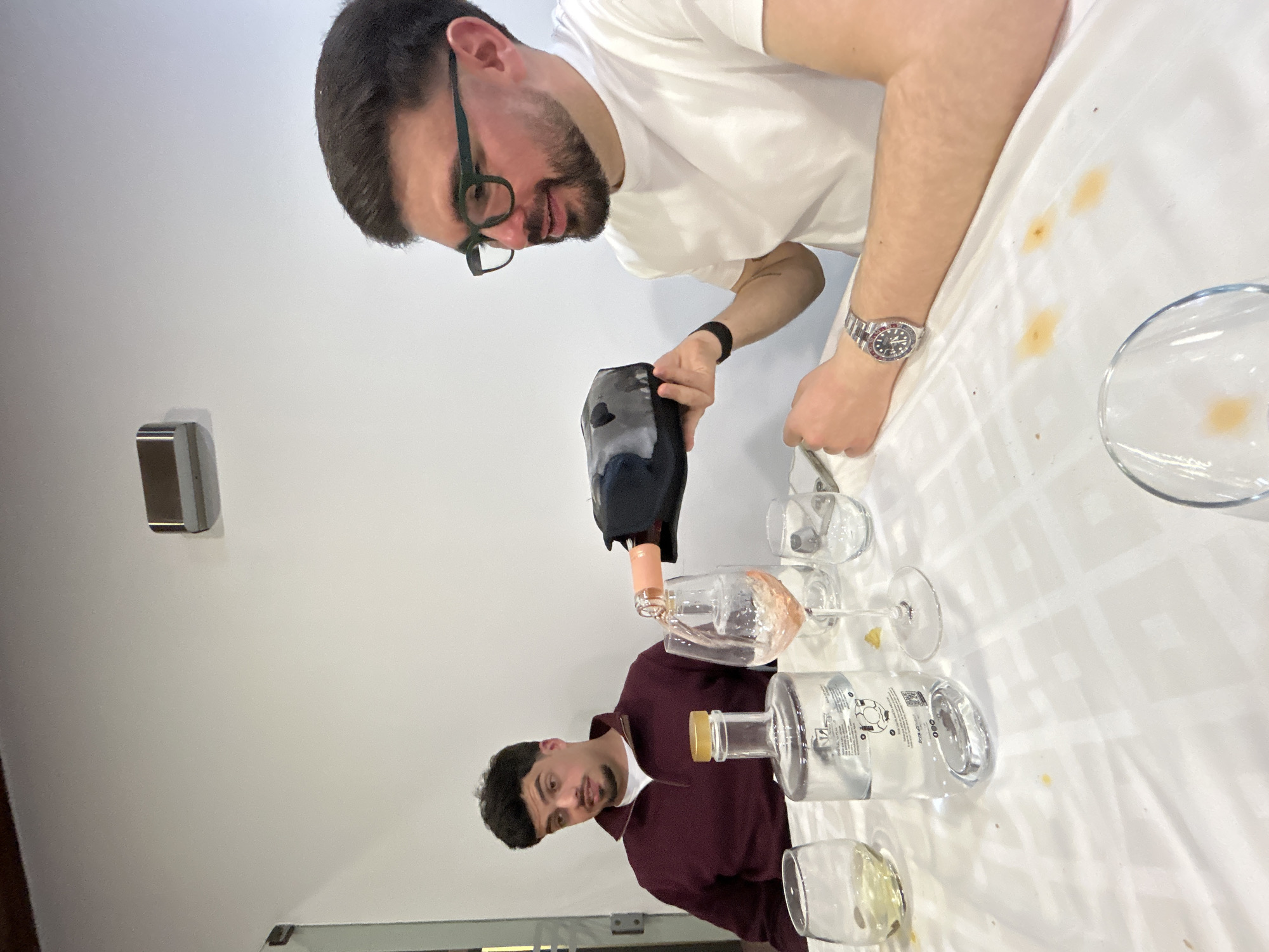 Two men are interacting around a table covered with a white tablecloth. The man at the top, wearing glasses and a white t-shirt, is pouring a liquid from a black container into a glass. The man at the bottom, wearing a burgundy top, is observing. Several glasses and bottles are on the table, some containing liquid. The location appears to be an indoor space with white walls. The city of Mirandela, Portugal is not visible in the image.