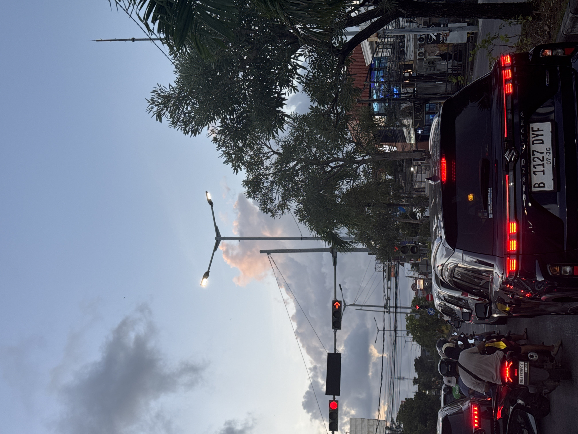 Here is a detailed description of the image:
The image shows a busy street scene, likely in a city in Indonesia, based on the license plates. The perspective is from within or beside a line of stopped vehicles at a traffic light. The traffic light is showing a red signal, and there are numerous motorcycles and at least three cars in the frame. The license plate of the car closest to the camera reads "B 1127 DYF." Other visible license plates are partially obscured, but one reads "DK6869FCY". The cars are dark in color.
Overhead are trees, power lines, street lights, and a complex of buildings. The sky is mostly light blue with some clouds.
The image suggests heavy traffic congestion.