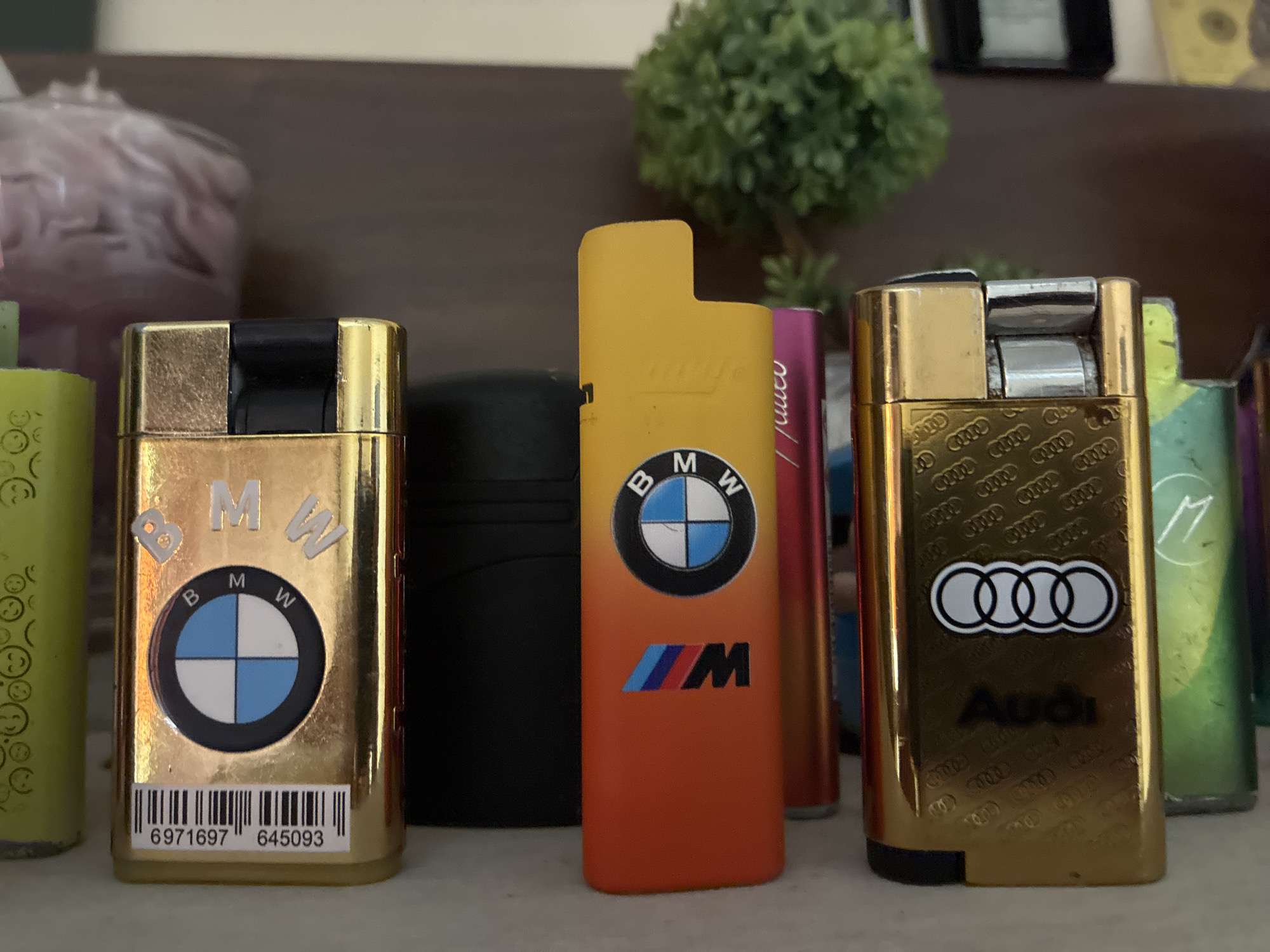 The image displays a collection of lighters, likely on a shelf or tabletop, in Varna, Bulgaria. In the foreground, three lighters are prominently featured.
On the left, a gold lighter showcases the BMW brand. It features raised silver "BMW" text and the classic BMW roundel logo in blue, white, and black. Below the logo, a white sticker with a black barcode is visible, containing the numbers "6971697 645093". The top part of this lighter is black, seamlessly transitioning into gold.
The central lighter has a striking orange to yellow gradient. It also features the BMW roundel logo and, beneath it, the BMW M logo with its characteristic blue, dark blue, and red stripes alongside a black "M". Subtle, stylized text, possibly "BMW", is faintly visible on its yellow upper section.
To the right, a gold Audi-branded lighter is shown. It prominently displays the Audi rings logo with silver outlines and a black fill, with the word "Audi" printed in black beneath it. The gold surface of this lighter is patterned with numerous smaller, embossed or printed Audi rings. This lighter, along with the left gold BMW one, shows some signs of wear.
Several other lighters are partially visible in the scene. On the far left, a green lighter with a pattern of smiley faces can be seen. Behind the central BMW lighter, a black, cylindrical object is partially obscured. Further right, a pink or purple lighter is partly visible, and another green lighter is present on the far right edge of the frame. In the background, a small, bushy green plant, likely artificial, sits on a shelf or surface, contributing to an indoor setting. The items are stationary, suggesting a display.