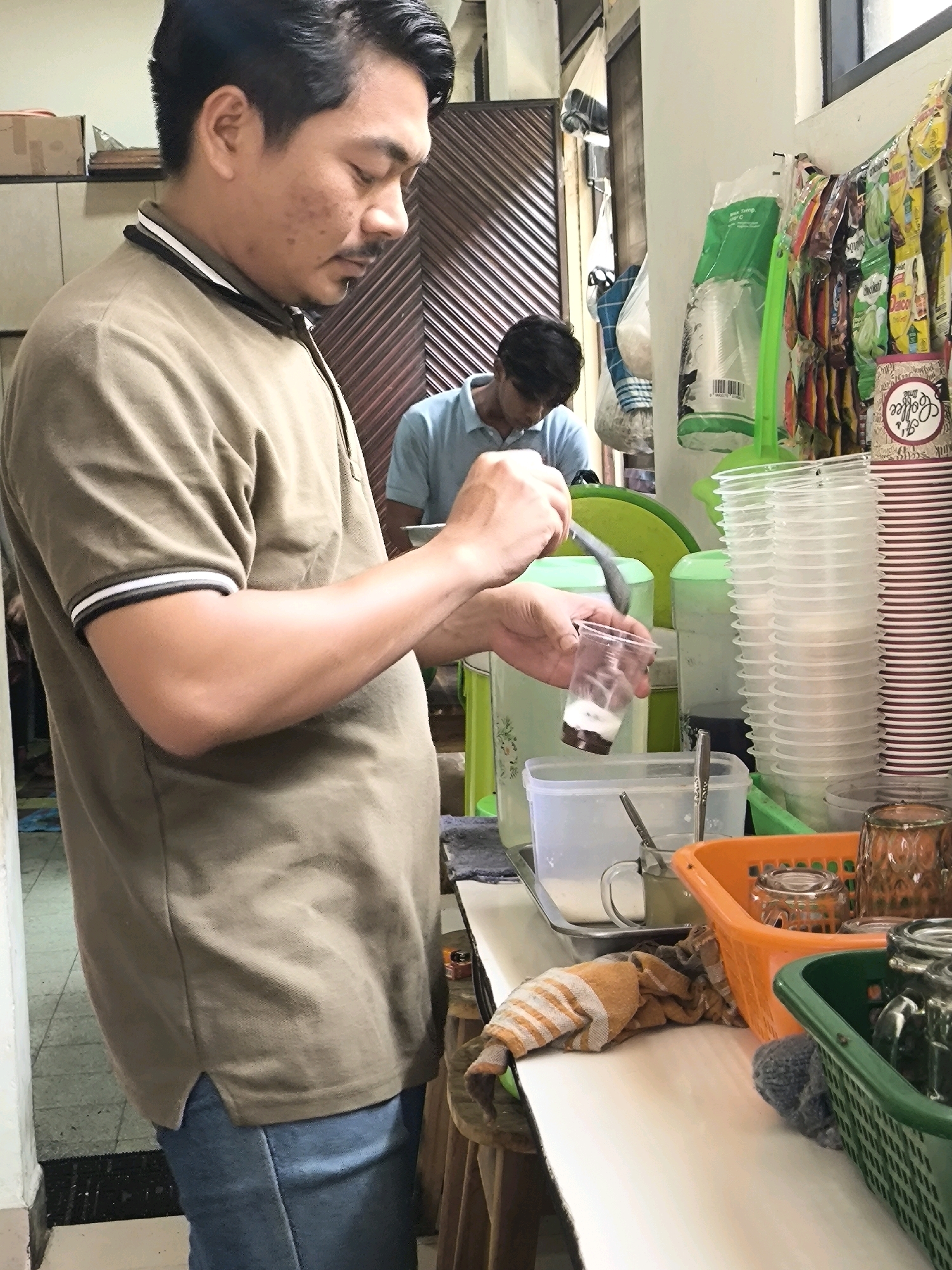 The image captures a scene inside a food stall in West Jakarta, Indonesia. A man in a light brown t-shirt with a striped collar is in the foreground, pouring something from a spoon into a clear plastic cup. He appears to be in the process of preparing a drink. Behind him, a younger man in a light blue shirt is visible, also looking down at something. The stall is cluttered with various items, including stacks of disposable plastic cups, a large green container, and an orange basket holding glass tumblers. Packaged snacks and drinks are displayed on a rack along the wall. The environment suggests a busy, perhaps small-scale food service operation.