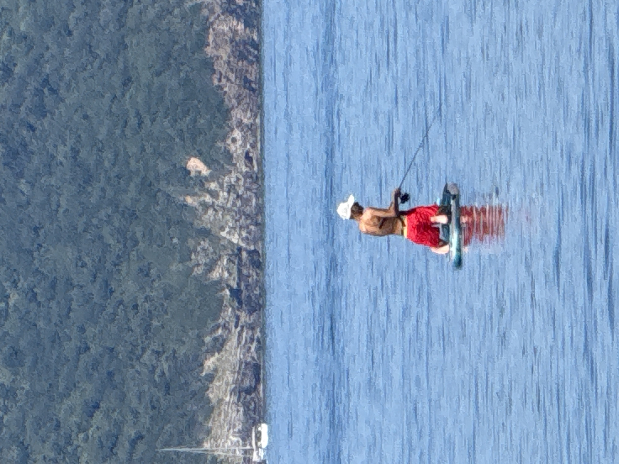 The media file shows a person kitesurfing on a bright, sunny day. The kitesurfer is a male wearing a white hat and red shorts, with his back to the camera. He is holding onto a kite string, preparing to jump or perform a trick on his board. The water is a clear, vibrant blue, reflecting the bright sky. In the background, a lush, green mountainside slopes down towards the water, dotted with some rocky outcrops and scattered buildings. A few boats are visible in the distance near the shore, indicating a coastal setting. The overall scene is serene and evokes a sense of outdoor recreation and enjoyment. The bright sunlight suggests it is midday.