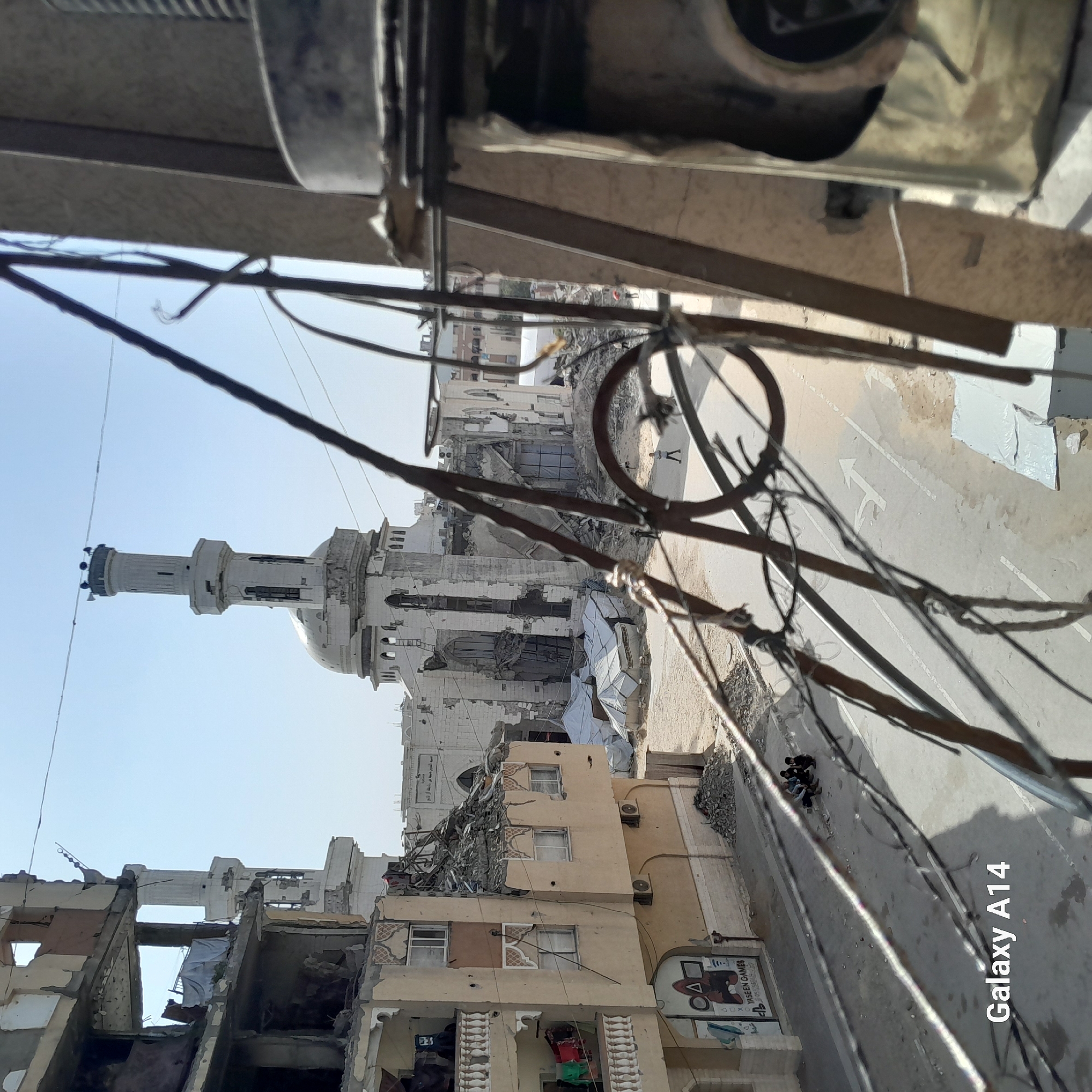The image, viewed in its upright orientation, depicts a scene of extensive destruction in an urban environment, likely a residential or commercial district. In the mid-ground, a prominent white mosque with a tall minaret and a dome is heavily damaged, with large sections of its structure missing and walls crumbling. Surrounding the mosque are several other multi-story buildings, also showing significant damage, with exposed interiors, shattered windows, and debris littering the ground.
In the immediate foreground, a dense network of tangled wires and what appear to be parts of an overhead structure or balcony obscure the top and left portions of the view. A dusty street runs through the scene, featuring faded white road markings, including directional arrows. A small group of approximately three individuals is visible on the street in the mid-distance, with another solitary figure further down the road.
To the right, a partially collapsed yellow building features air conditioning units mounted on its exterior. At its base, a commercial sign is visible, displaying "YASEEN GAMES" and "PS4," along with a logo resembling the symbols from the "Squid Game" series (circle, triangle, square). A white truck or van with an indistinct graphic on its side is parked below this building.
The sky is clear and bright blue, suggesting daytime, possibly morning or early afternoon, with shadows indicating a strong sun. The overall scene conveys a powerful sense of devastation and the aftermath of a major destructive event, such as conflict or disaster. A "Galaxy A14" watermark is visible in the bottom right corner of the image.