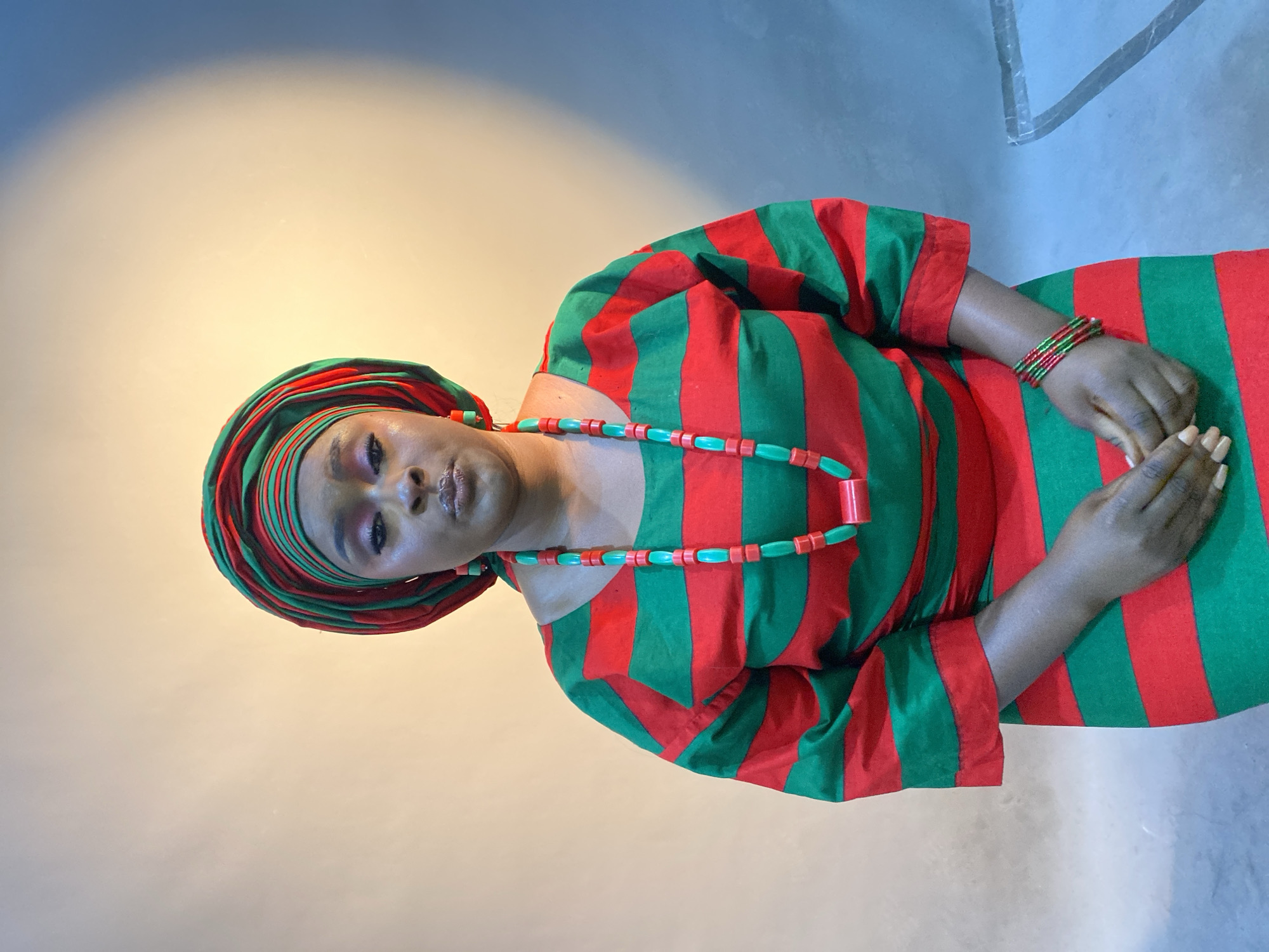 A woman is shown in an eye-level portrait. She is adorned in a vibrant, striped garment of red and green. Around her head, she wears a matching headwrap in the same colors. A necklace of alternating red and green beads rests around her neck. Her wrists feature multiple bangles in coordinating shades. Her hands are gently clasped in front of her. The background is a light gradient of cream and light grey. The location is Gura Top, Nigeria.