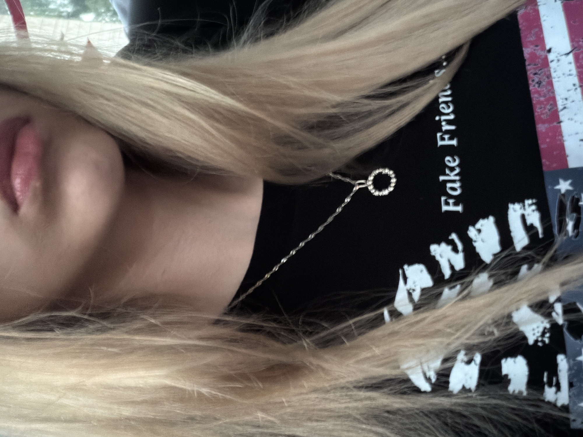 The image shows a close-up of a person with blonde hair and fair skin. The person is wearing a black t-shirt with white text that says "Fake Friends...". A silver necklace with a circular pendant adorned with small stones is visible on the person's neck. The background is out of focus and appears to be outdoors with some greenery. The bottom portion of the t-shirt displays a distressed American flag pattern.