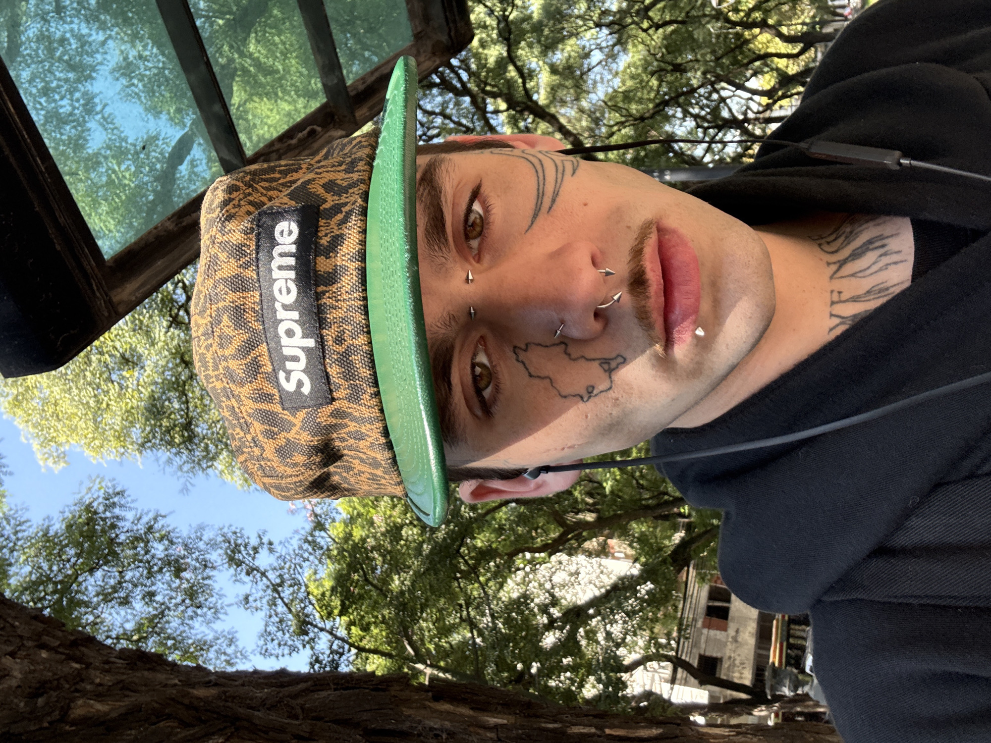 A person is wearing a Supreme brand baseball cap, with a green brim and a black and brown animal print design. The person has multiple facial piercings, including piercings on the nose, lips, and eyebrows. They also have facial tattoos, and tattoos on their neck. They are wearing a black hoodie. The background shows green trees, blue sky, and some architecture that might suggest an urban park setting in Buenos Aires, Argentina.