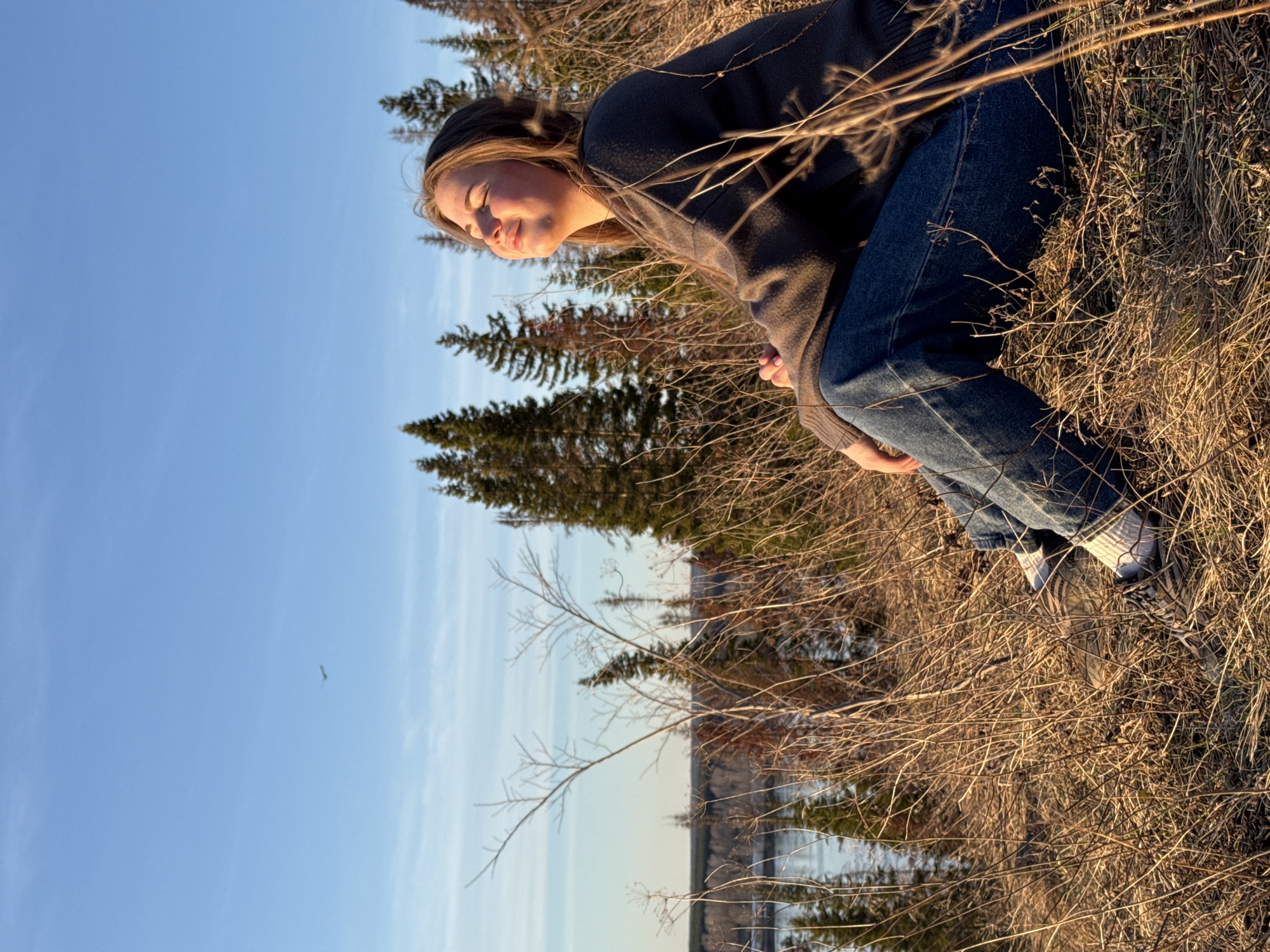 A young woman is sitting on a grassy hill in what appears to be a natural environment. She is wearing a dark sweater and blue jeans, with white sneakers. The backdrop includes pine trees and a body of water in the distance, possibly a lake or river. The sky is clear blue. Given the mention of Ольховка, Russia, it is plausible that this scene is located near that city, although specific landmarks are not discernible in the image to confirm the exact location. The photo appears to be taken during golden hour, based on the warm, low angle of the sun.