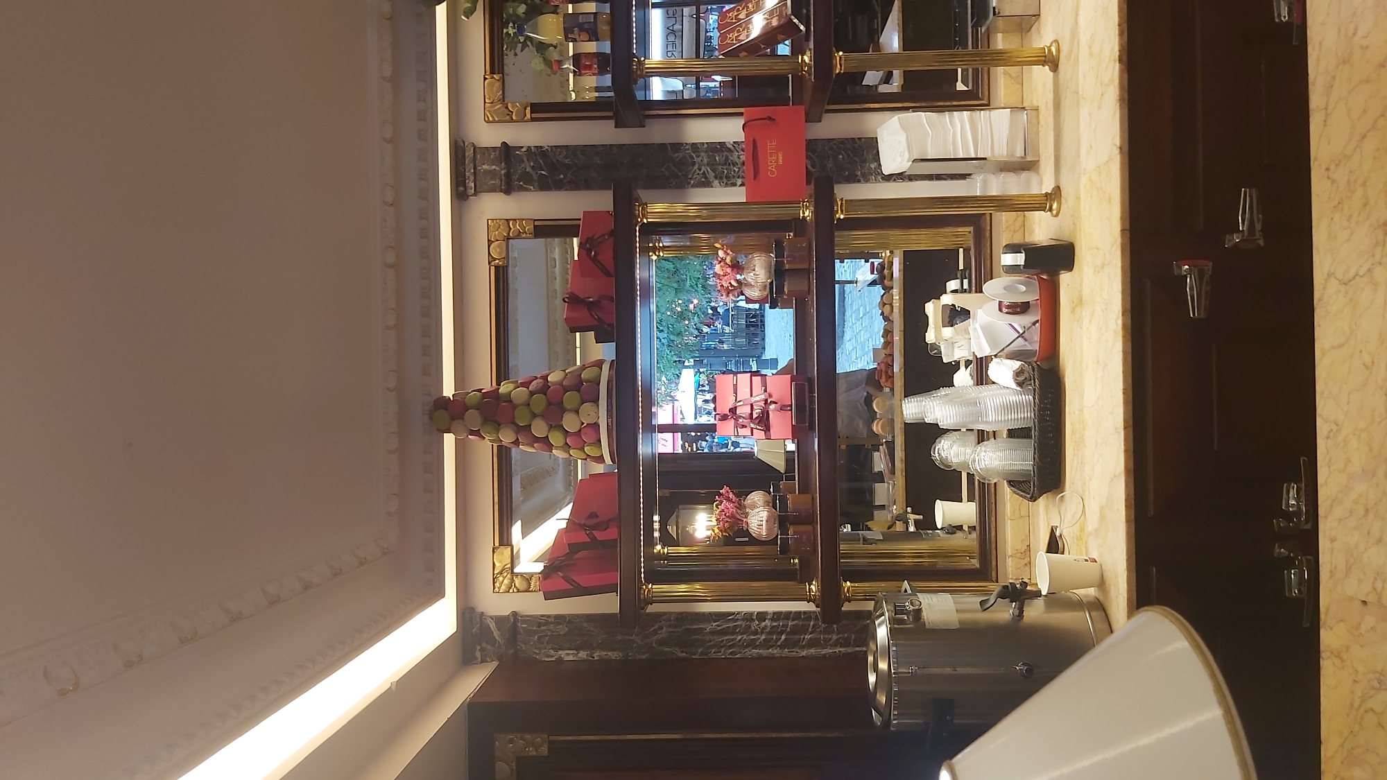 The image shows the interior of a patisserie in Paris, France. On display are various pastries, including a large croquembouche tower topped with macarons. Red gift boxes are arranged on shelves, and bottles of what appear to be juices or sodas are visible. A coffee or hot water dispenser is on the counter, along with disposable cups and other service items. Reflections in the mirrors behind the display show an outdoor scene with people and seating. The overall ambiance suggests a high-end establishment, possibly Carette, as indicated by a red bag with the brand name.