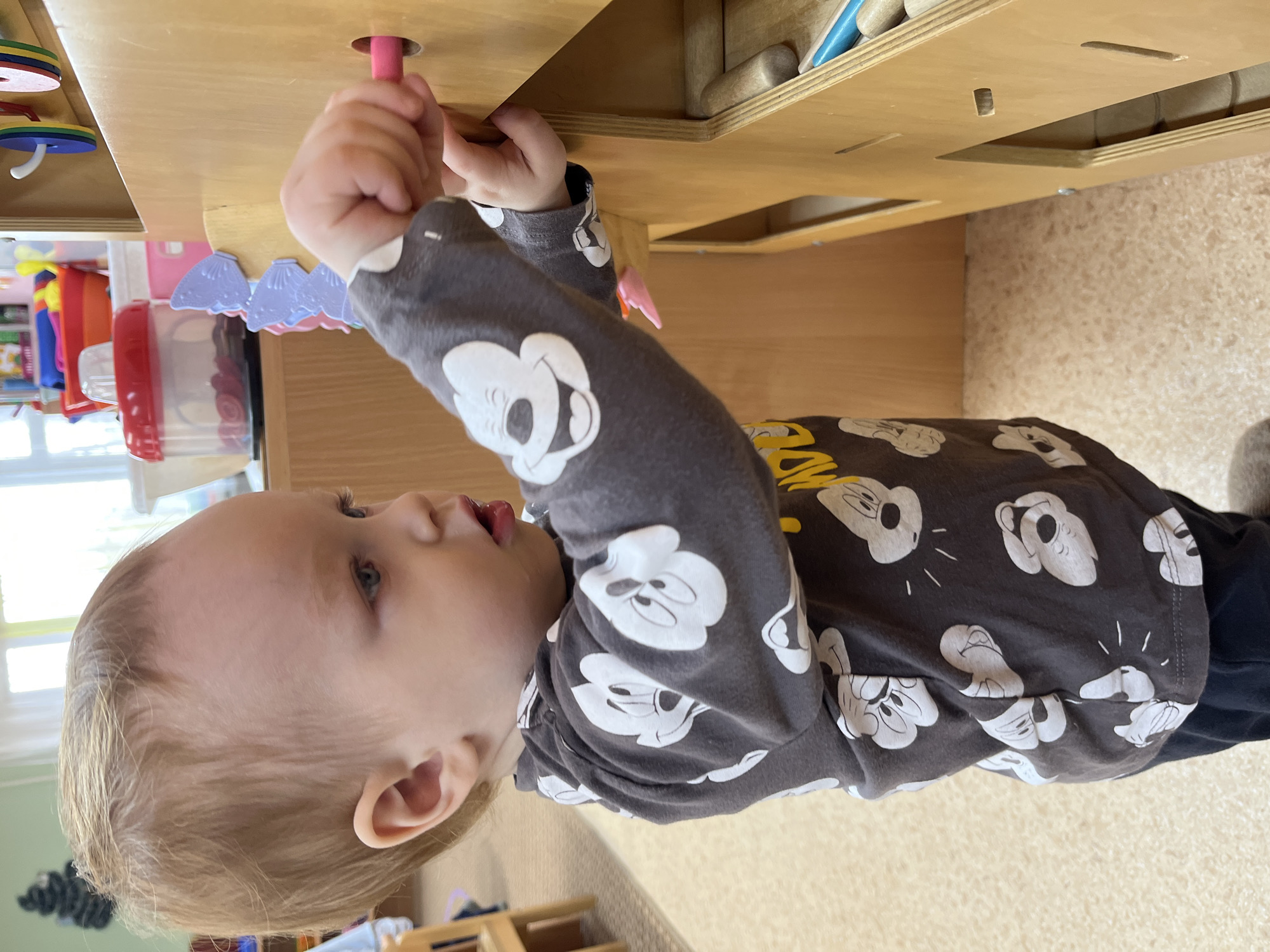 A young child wearing a long-sleeved gray shirt with white Mickey Mouse designs is playing with a wooden toy. The child is inserting a pink peg into a hole in the toy. Other colorful toys and items are visible in the background. The location is unknown, and there is no evidence to suggest that this image was taken in Chaikovsky, Russia.