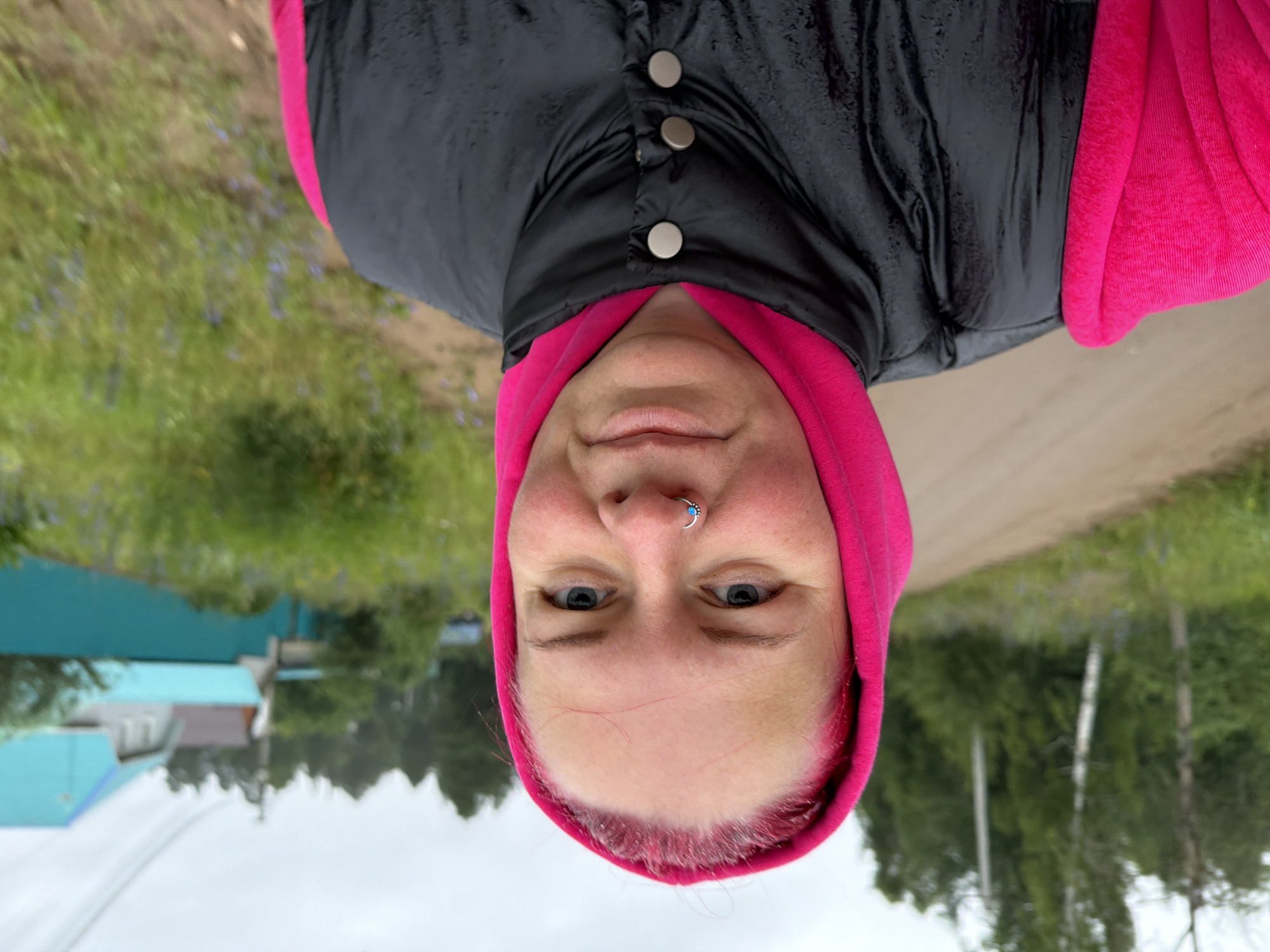 A close-up, upside-down, selfie-style photo shows a person wearing a pink hooded sweatshirt and a black puffer vest over it. The person has blue eyes, light skin, and visible nose jewelry. Their hair is pink-toned. The background is slightly blurred, showing a dirt road on the right and green vegetation on the left, with some distant houses and trees. The sky appears overcast, suggesting cloudy weather. The image seems to be taken outdoors in a rural or semi-rural setting, possibly in Chaikovsky, Russia, given the location context. The overall impression is casual and unposed.