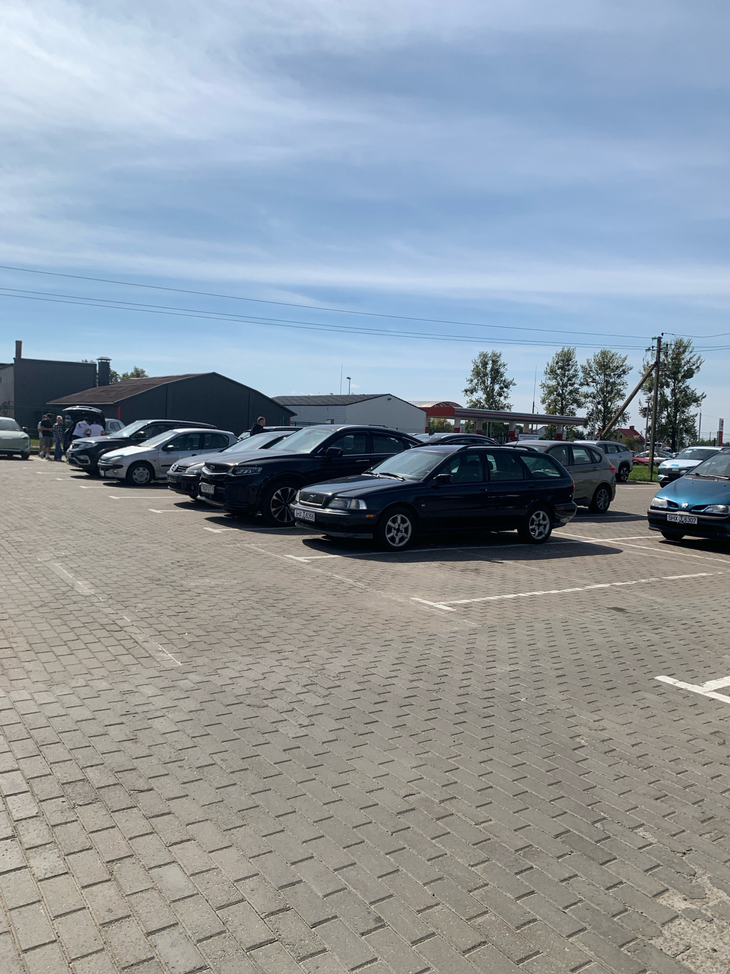 A group of cars are parked in a paved lot under a bright, cloudy sky. The lot is made of rectangular bricks laid in a herringbone pattern. In the background, a row of low-rise buildings with dark roofs are visible, along with several trees. A few people are standing near the cars in the left side of the frame. One of the cars in the foreground is a dark blue station wagon. Another dark-colored SUV is parked to its right, with a white license plate with black lettering. A light-colored car is parked to the left of the station wagon, and a silver sedan is parked to its left. The scene appears to be a parking lot in an industrial or commercial area, possibly near a gas station visible in the mid-ground. The overall atmosphere is one of a typical daytime scene in an outdoor urban or semi-urban environment.