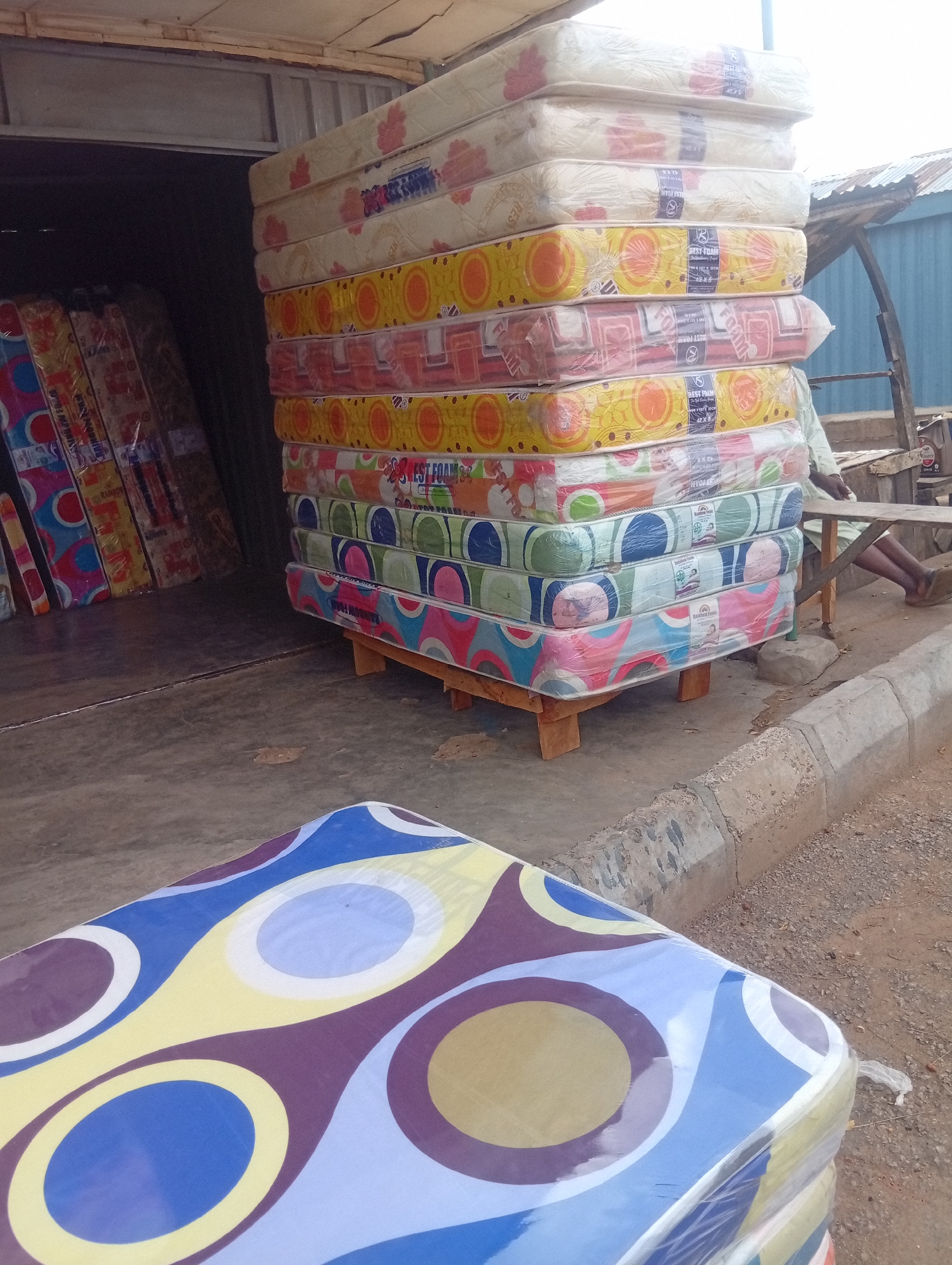 The image shows a stack of mattresses for sale in Birnin Kudu, Nigeria. The mattresses are of varying colors and patterns, and are wrapped in plastic. The stack is placed on a wooden pallet near what appears to be a store or warehouse, with additional mattresses visible inside. In the foreground is another stack of mattresses with a blue, yellow, and purple circle pattern. A person is sitting at a table in the background. The location is a street or sidewalk next to the building.