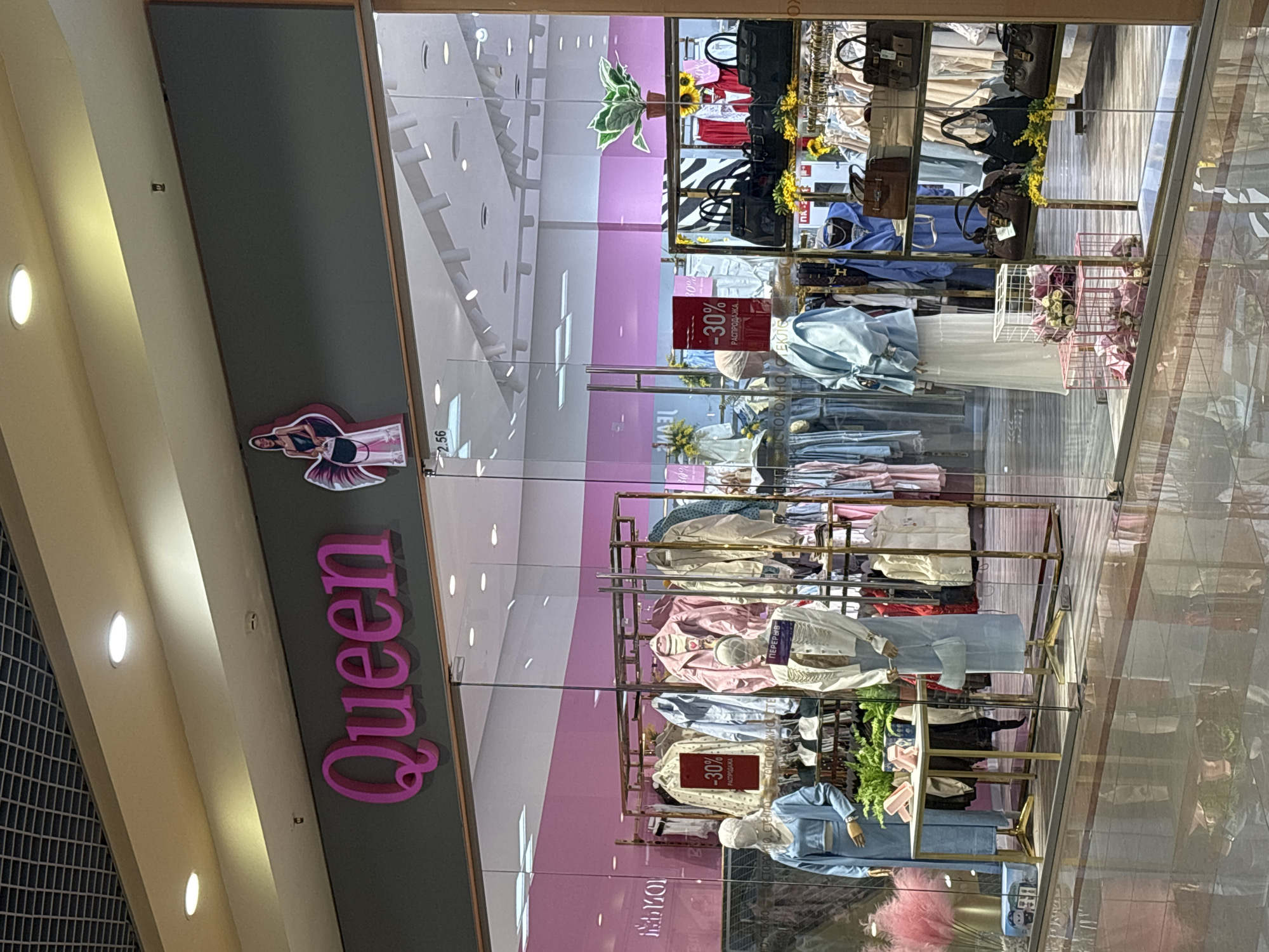 The image shows the storefront of a clothing store named "Queen" located in Ivanovo, Russia. The store features a large glass window displaying mannequins dressed in women's clothing, as well as racks of clothing and handbags. A prominent sign on the window indicates a "30% SALE." The store's name, "Queen," is displayed in large pink letters above the window. The store appears to be located within a shopping mall, as evidenced by the ceiling grid and lighting. The floor is reflective, showing a mirror image of the store's interior.