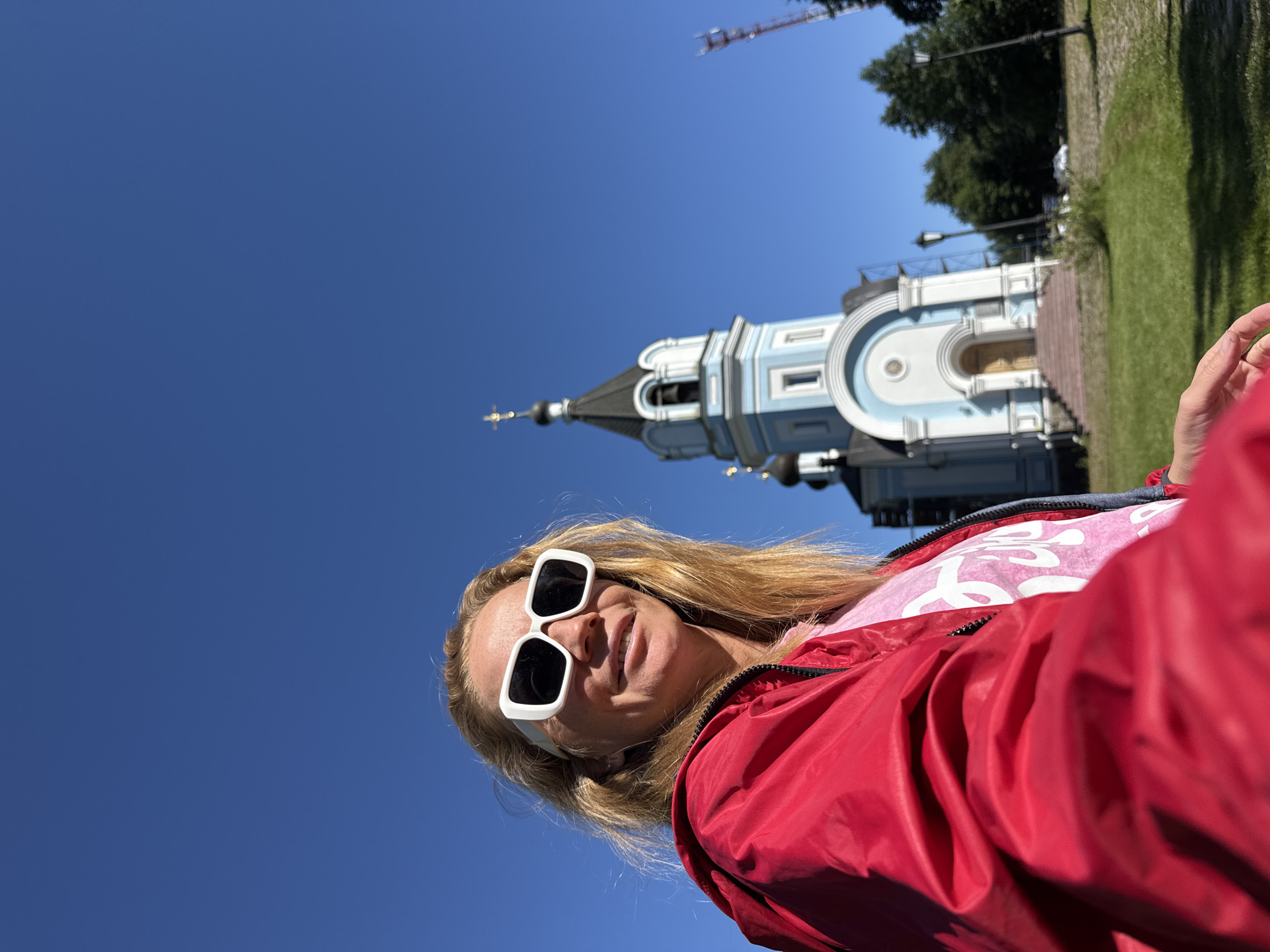 In St. Ozerki, Russia, a woman with blonde hair and white-framed sunglasses is captured in a selfie. She is wearing a vibrant red jacket over a pink t-shirt. Behind her, a striking blue and white Orthodox church with a black dome and golden crosses stands against a clear blue sky. The church has a prominent bell tower and a grand entrance with stairs. Surrounding the church, a green lawn is dotted with trees and lampposts, and a telecommunication tower is visible in the distance. The woman is positioned in the foreground, with her face slightly turned towards the camera and a slight smile. The image is taken outdoors on a bright, sunny day.