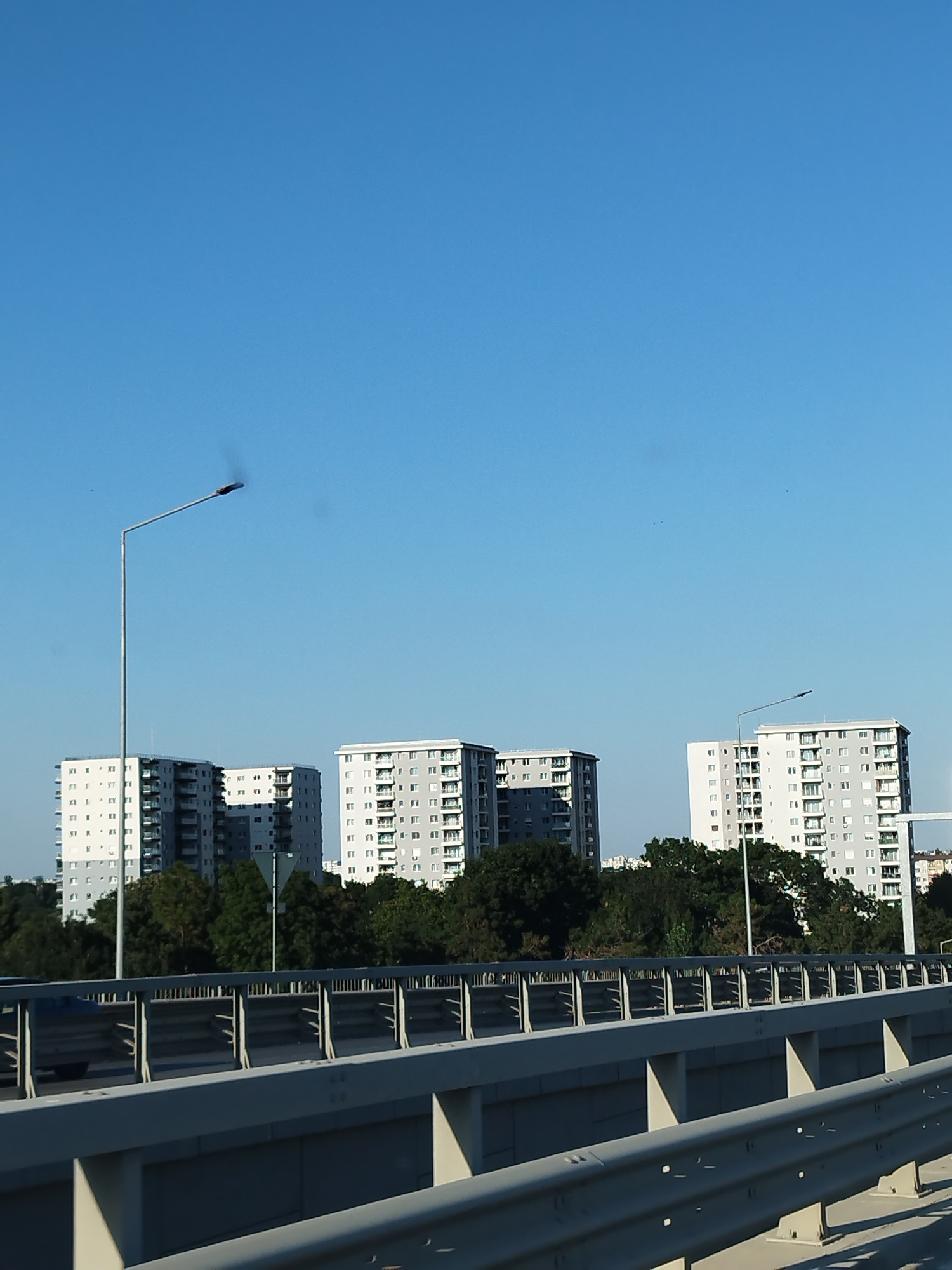 In Bragadiru, Romania, on a clear day, a view from a highway overpass reveals several tall, modern apartment buildings in the background. The buildings are primarily light gray and white, with numerous windows and balconies. Trees form a green line in front of the buildings. A tall street lamp stands to the left, its arm extending over the road. In the foreground, a concrete barrier and guardrails of the highway are visible, with a car driving on the road below. The sky is a vibrant, cloudless blue.