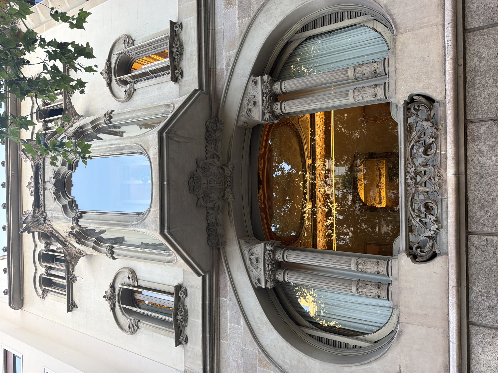 The image shows the facade of Casa Amatller, a modernist building located on Passeig de Gràcia in Barcelona, Spain. The photograph is taken from a low angle, looking upwards at the building's ornate architectural details.
The most prominent features are the large, arched windows with intricate stone carvings. The large oval window in the lower half of the image is framed by decorative columns and topped with a balcony with a wrought-iron railing. The window itself reflects the warm glow of interior lighting and the surrounding trees. Above this, a smaller, projecting bay window with a curved glass facade is visible, also adorned with sculptural elements. To the left and right of the central bay window are narrower, vertical windows with elaborately shaped frames.
The building's facade is a light cream color, contrasting with the darker stone of the lower levels. Lush green leaves from a tree overhang the top of the building on the right side of the frame, adding a natural element to the urban scene. The architectural style is clearly modernist, characterized by organic forms, detailed ornamentation, and a sense of fluidity in its design. The image captures a detailed view of the building's exterior, highlighting its unique and artistic construction. There are no people or animals visible in the image.