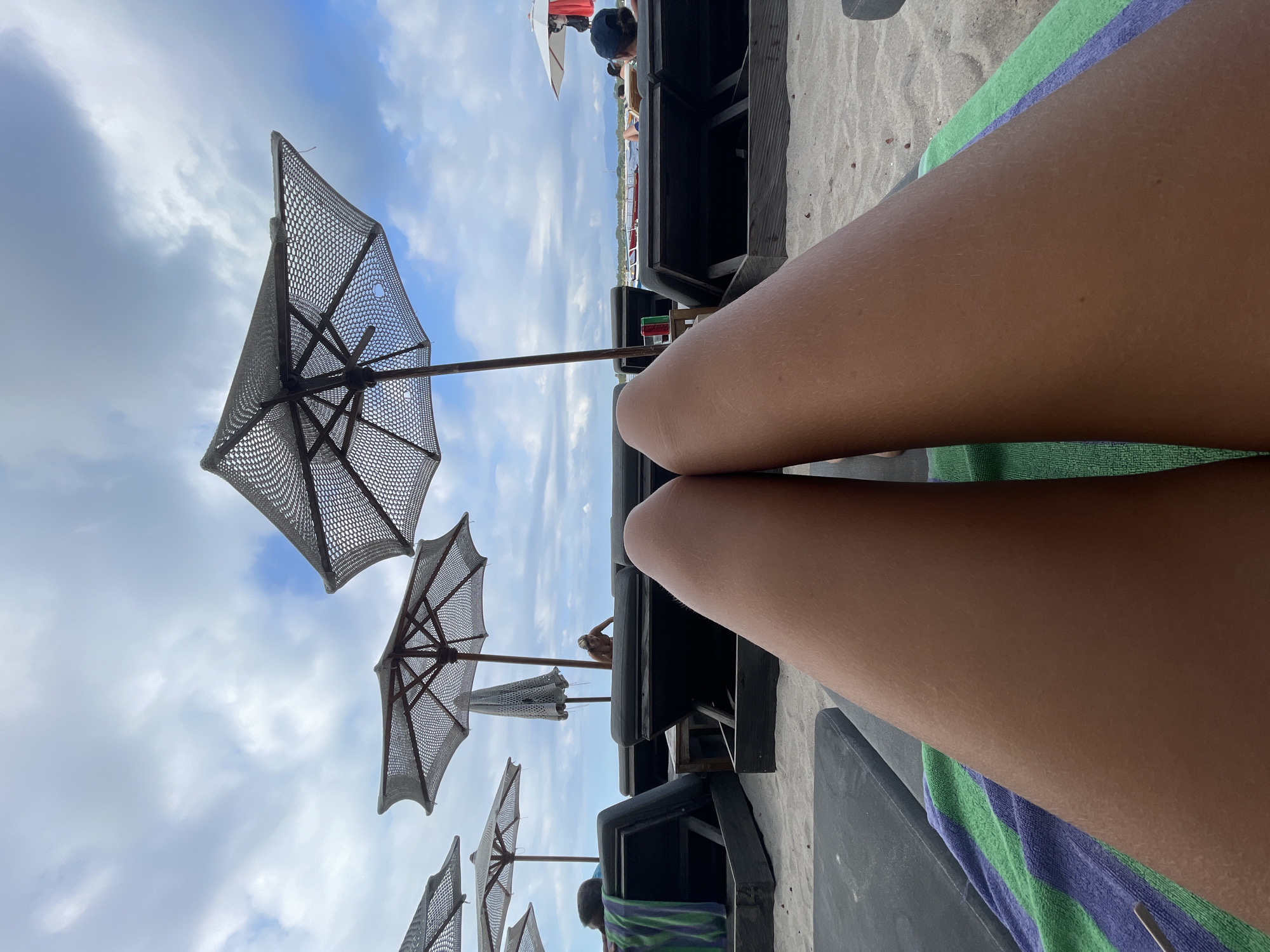 The image depicts a first-person perspective of a person relaxing on a beach lounge chair. The foreground is dominated by the person's legs and a striped towel. In the background, rows of empty lounge chairs and umbrellas are visible on a sandy beach. Several people are sparsely scattered throughout the background, some seated on lounge chairs and one standing near an umbrella. The sky above is cloudy, with patches of blue. The overall scene suggests a relaxed atmosphere on a beach. The location is identified as Unknown, Indonesia.