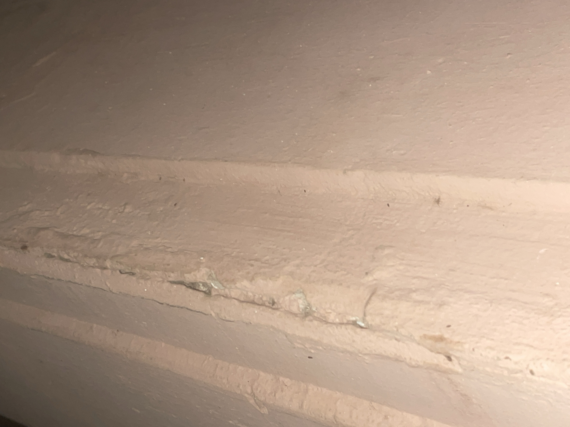 The image shows a close-up view of a textured, light pink painted surface, likely a wall or column. A distinct vertical seam or crack runs down the center of the image, with visible signs of damage and material loss along its edges. The texture of the paint is uneven, with some areas appearing smoother and others rougher. The lighting suggests an indoor environment, possibly with artificial light sources casting subtle shadows. There are no people or discernible objects other than the surface itself. The location is unspecified within Howayi, Nigeria.